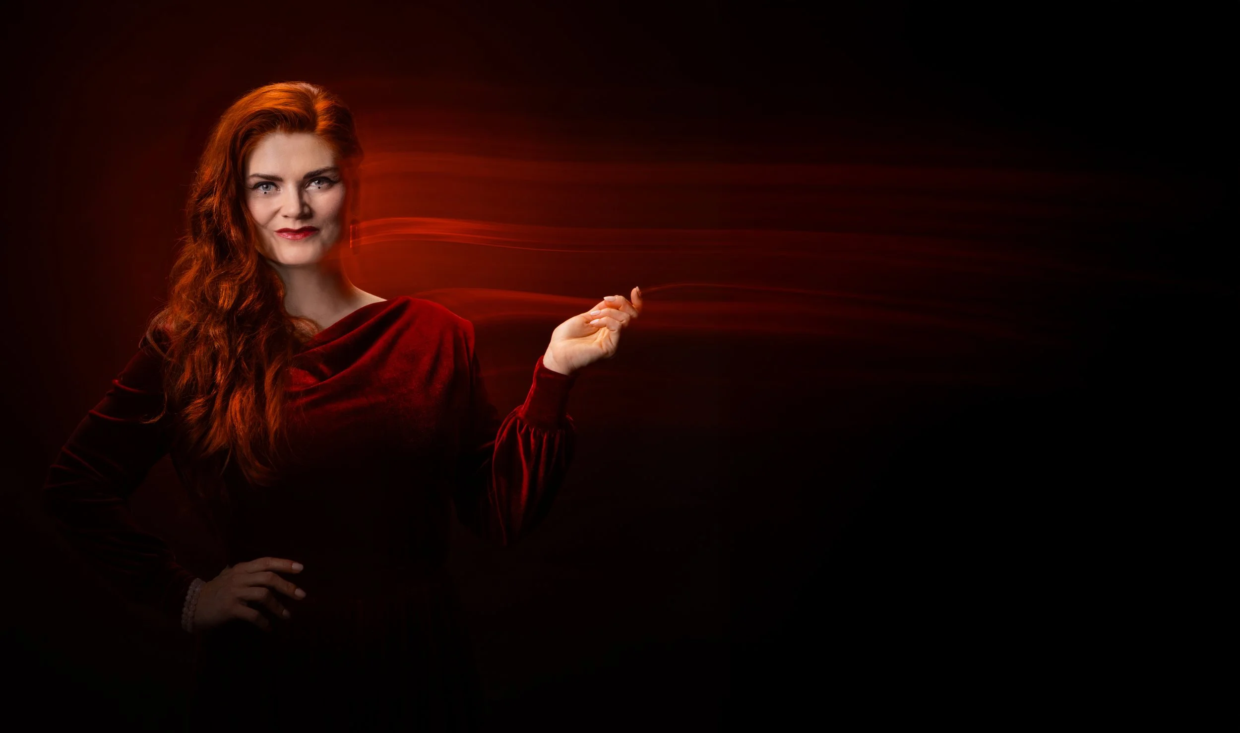 A woman with long red hair in a red velvet dress standing against a dark background with red light streaks emanating from her hand.