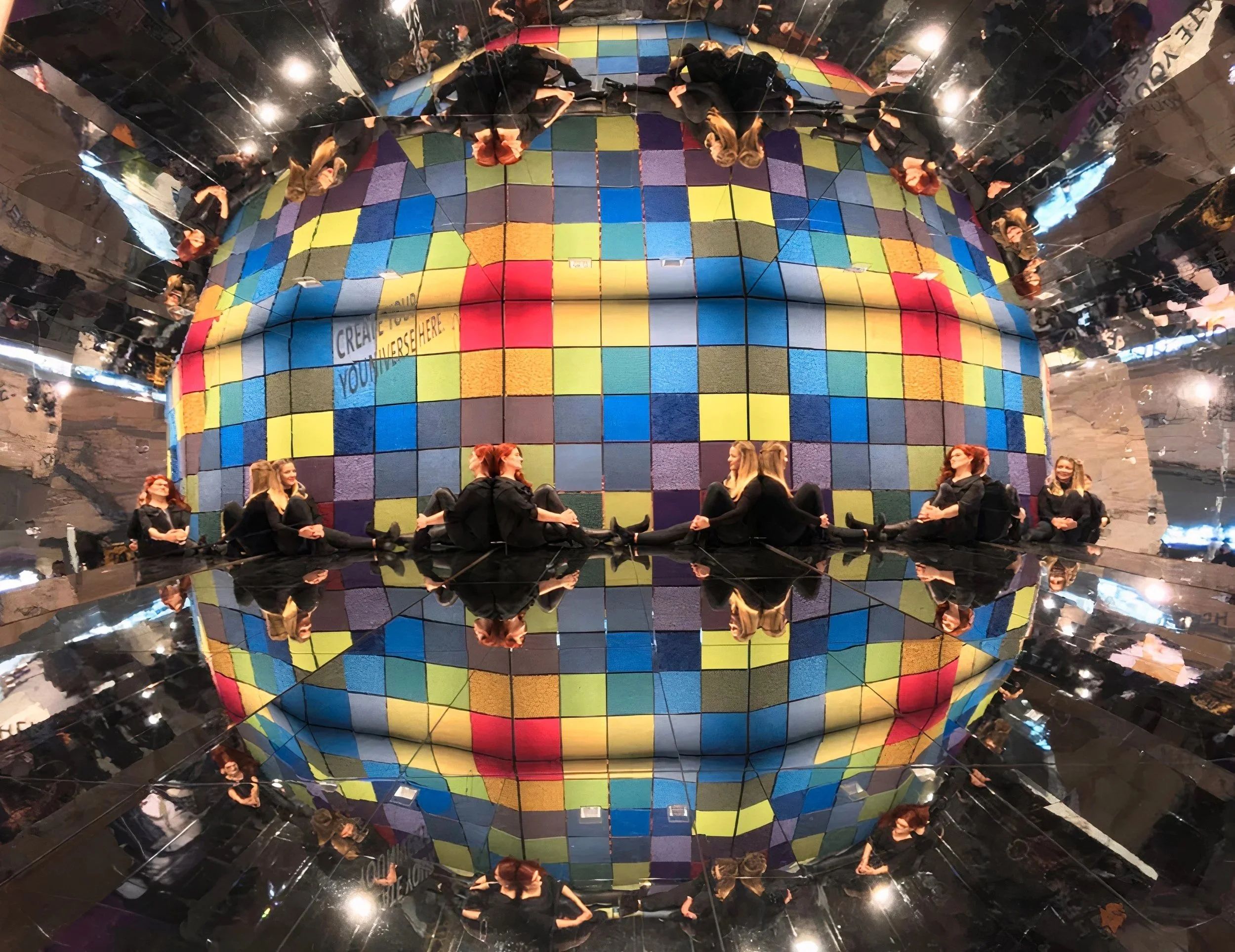 Multiple women sitting in a circle on the floor inside a mirrored room with a colorful, mosaic-like, spherical art installation.