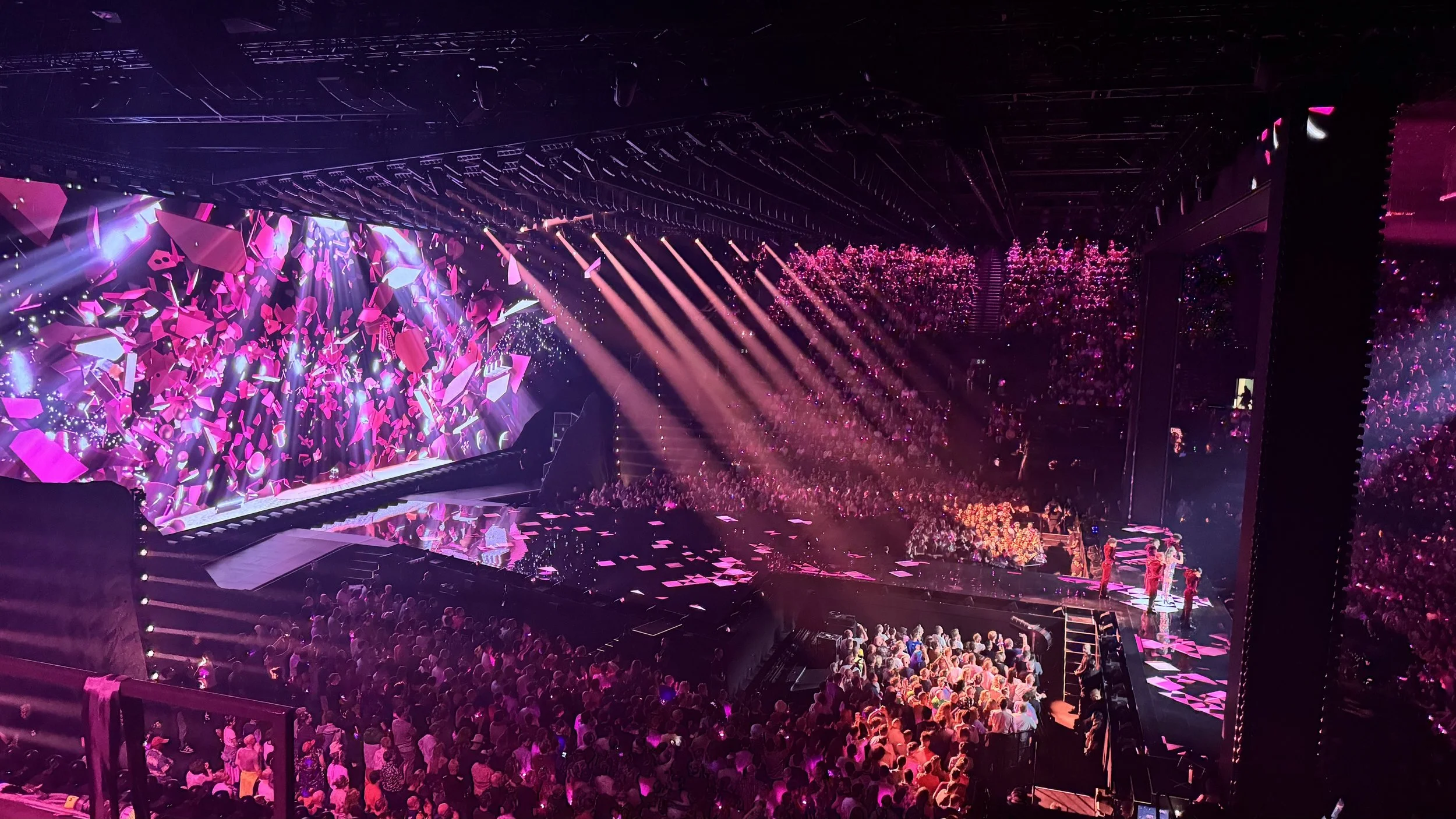 A concert or event venue with a large audience, illuminated by pink and purple stage lights, with a dynamic digital display on the stage featuring abstract shapes and designs.