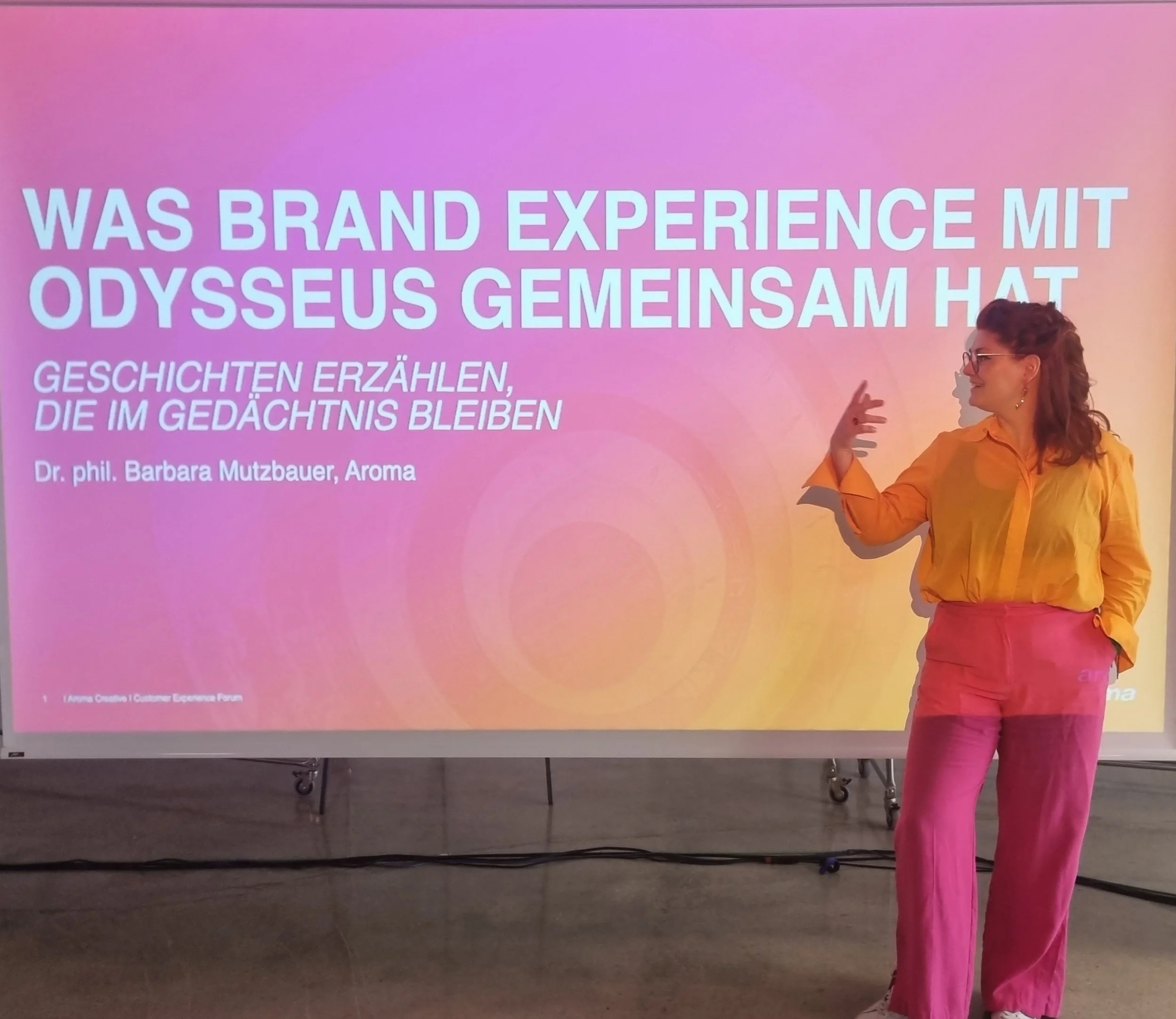 A woman in pink pants and a yellow shirt standing next to a presentation screen with a colorful gradient background. The screen displays text in German and English.