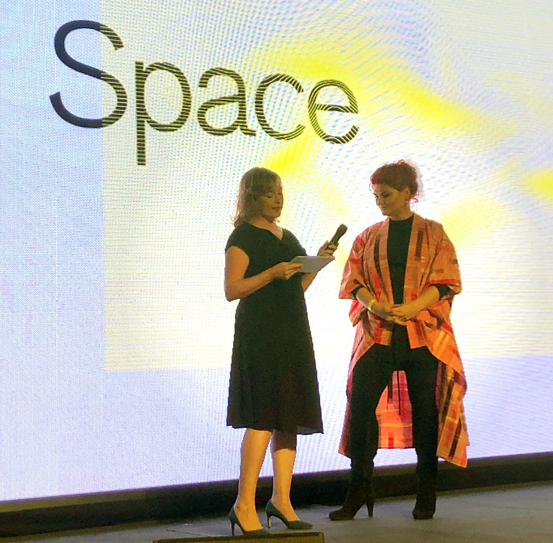 Two women on stage, one holding a microphone and paper, the other holding a notepad, standing in front of a large screen that says 'Space.'