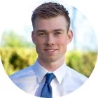 A young man with short brown hair smiling outdoors, wearing a light blue shirt and dark blue tie, with green trees in the background.