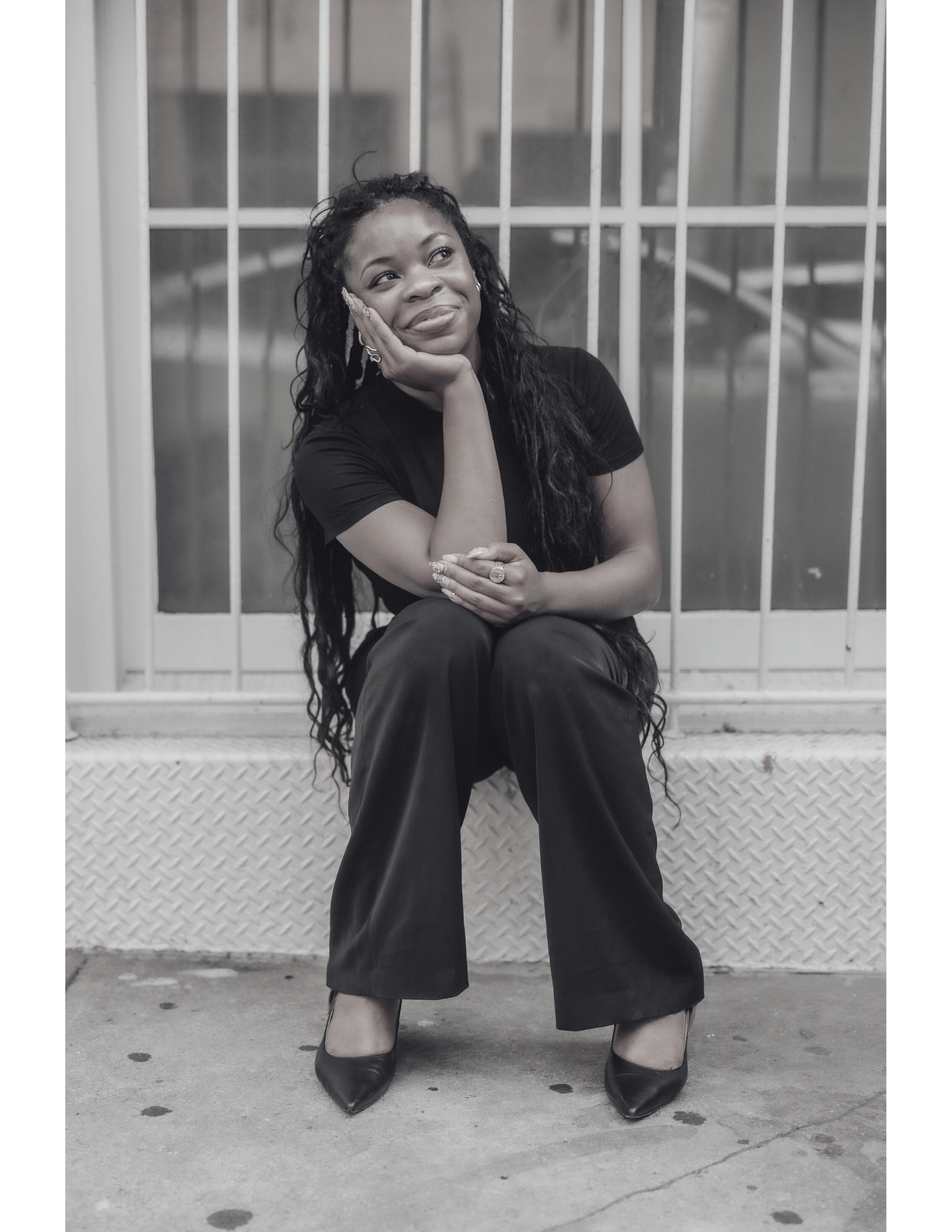 A woman with long curly hair sitting in front of a window with bars, resting her face on her hand, smiling thoughtfully, wearing a black t-shirt, black wide-leg pants, and black heels.