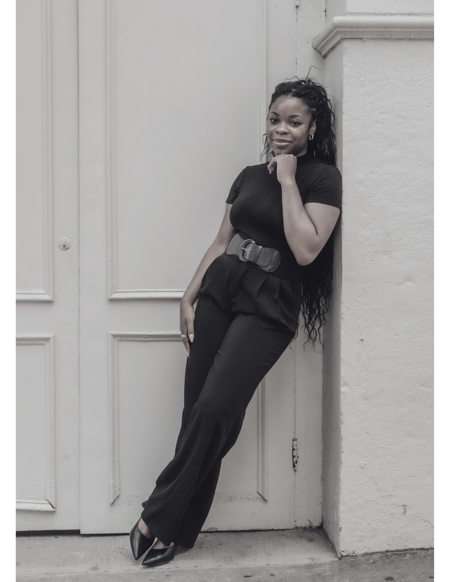 A woman standing against a door, wearing a black t-shirt, black high-waisted pants with a wide belt, and black heels, with one hand resting on her chin and the other on her thigh.