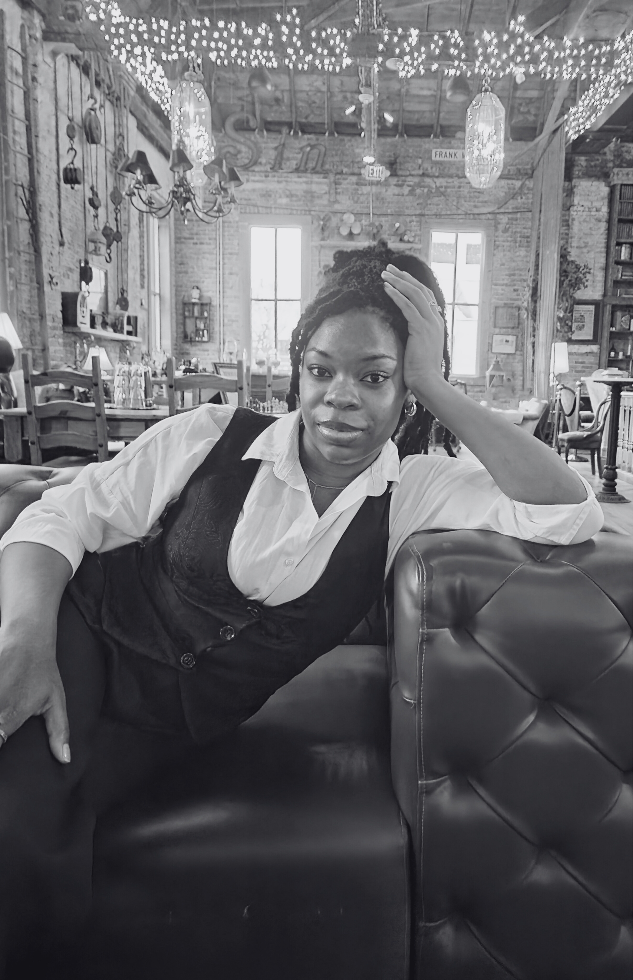 A Black woman with short dreadlocks, wearing a white shirt and black vest, relaxing on a leather couch with one arm resting on the armrest and her hand on her head, in a rustic, decorated indoor setting with string lights and large windows in the background.