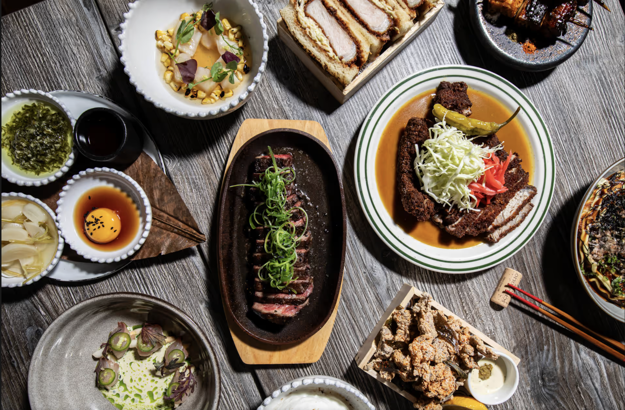 A variety of Asian dishes on a rustic wooden table, including sushi rolls, sliced beef with green onions, fried chicken, ramen, and various small side dishes with sauces and garnishes.