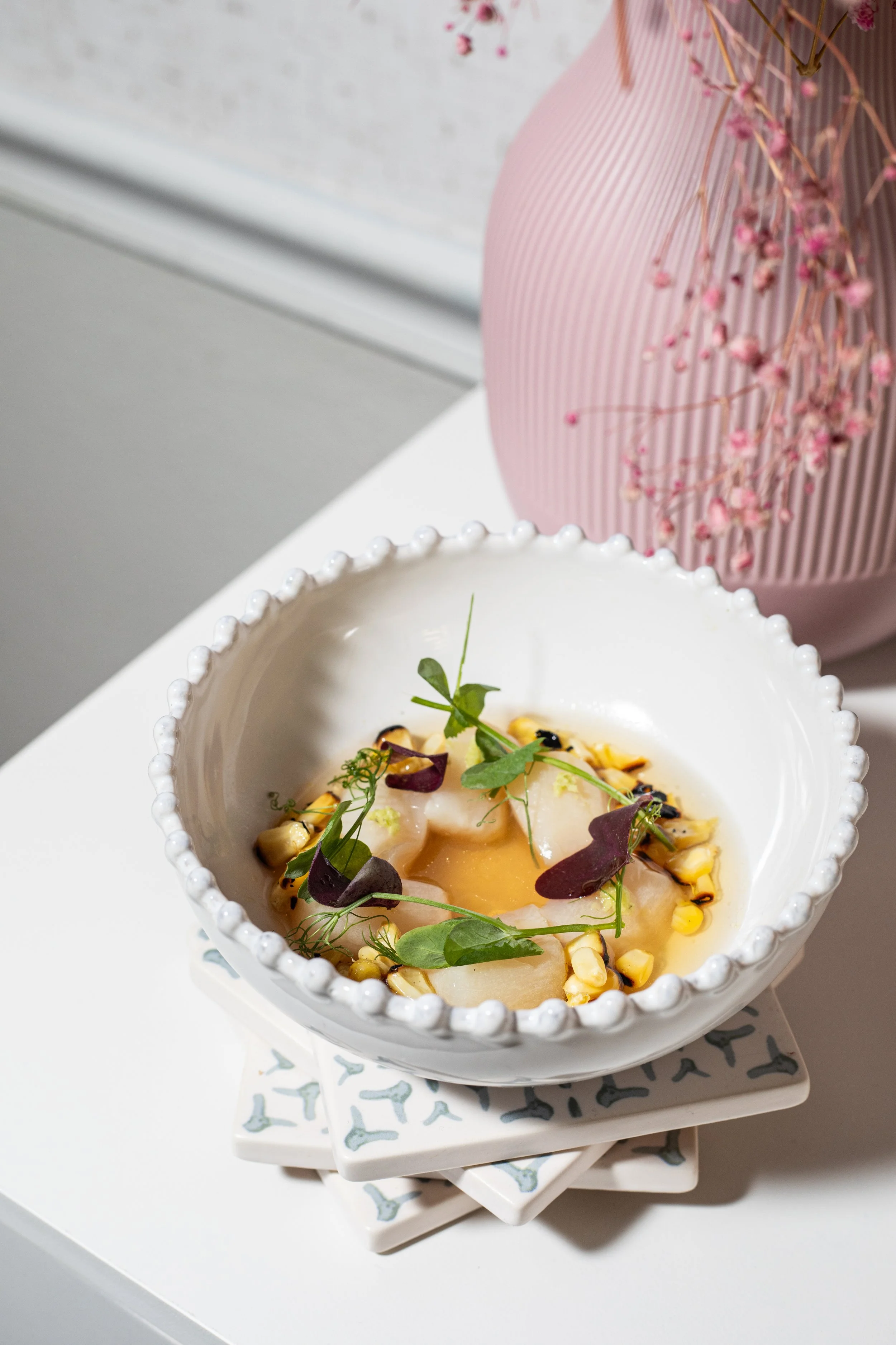 A white bowl with decorative edges containing a gourmet dish with a poached egg, microgreens, toasted corn, and edible flowers, placed on top of stacked patterned ceramic plates. A pink textured vase holding pink flowers is in the background.