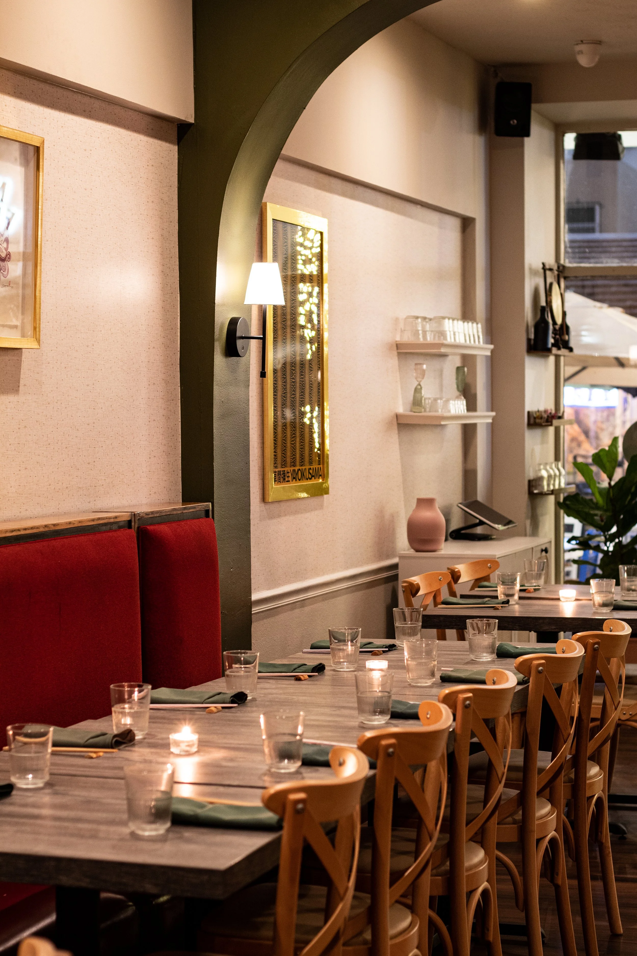 Interior of a restaurant with a long wooden table set with glasses, napkins, and candles, surrounded by wooden chairs, with wall-mounted shelves, artwork, and dim lighting creating a cozy atmosphere.