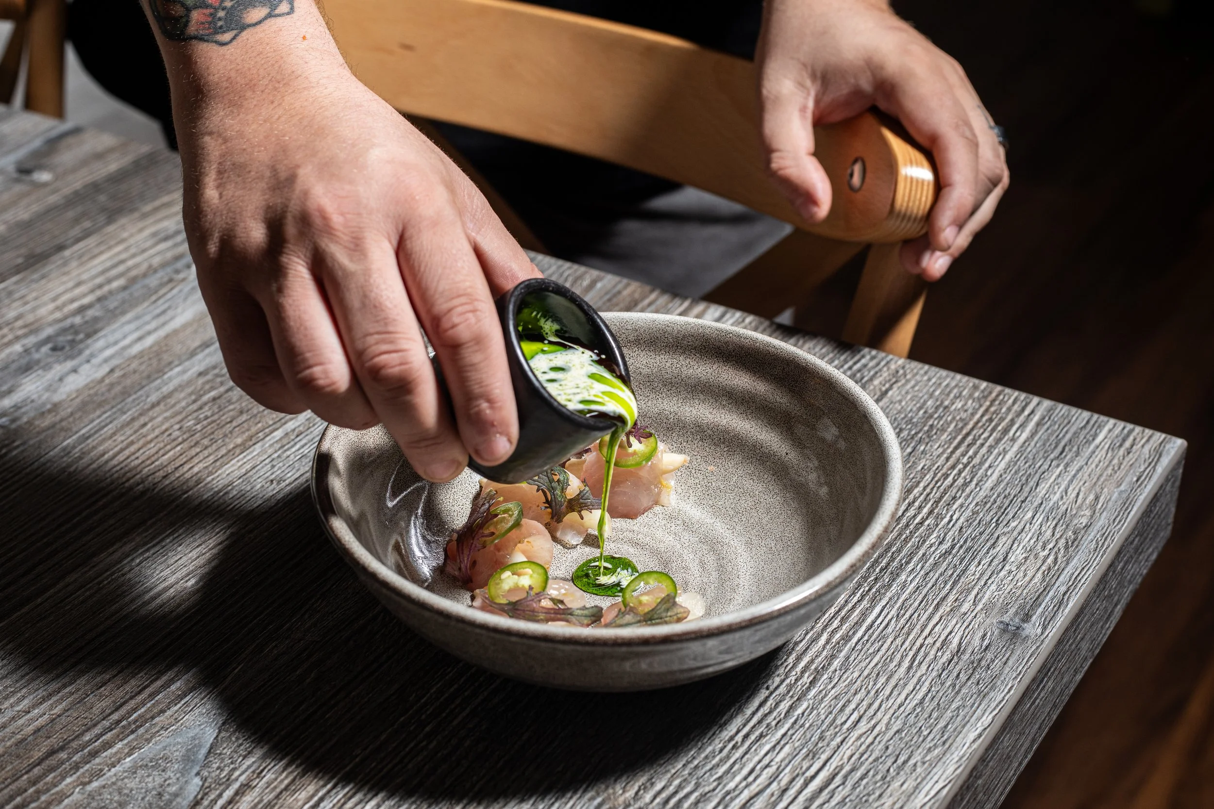 A chef pours green sauce over raw fish slices and sliced jalapeños in a grey ceramic bowl on a wooden table.