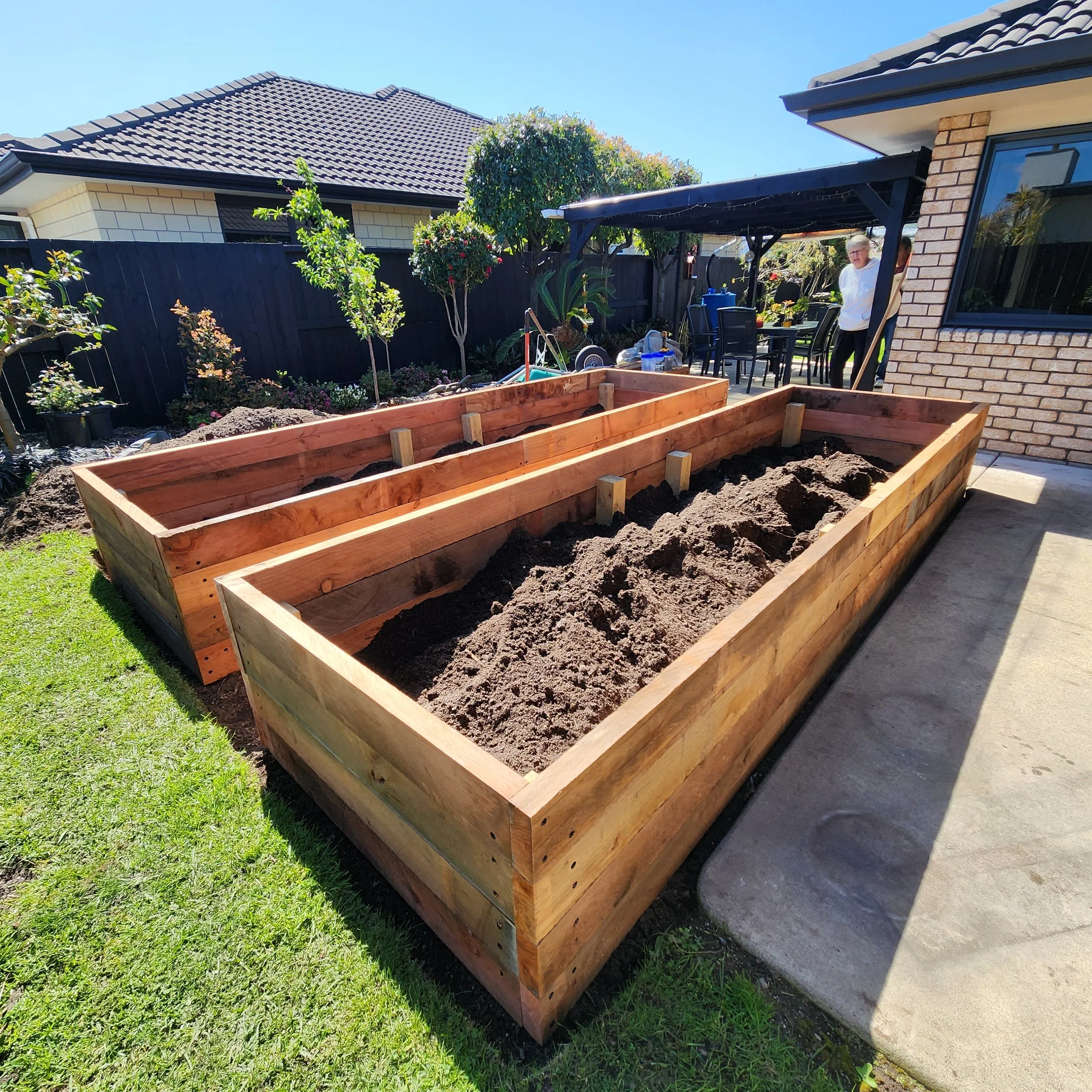 Two large wooden garden beds partially filled with soil in a backyard, with a house, plants, and a woman standing nearby under a patio in the background.