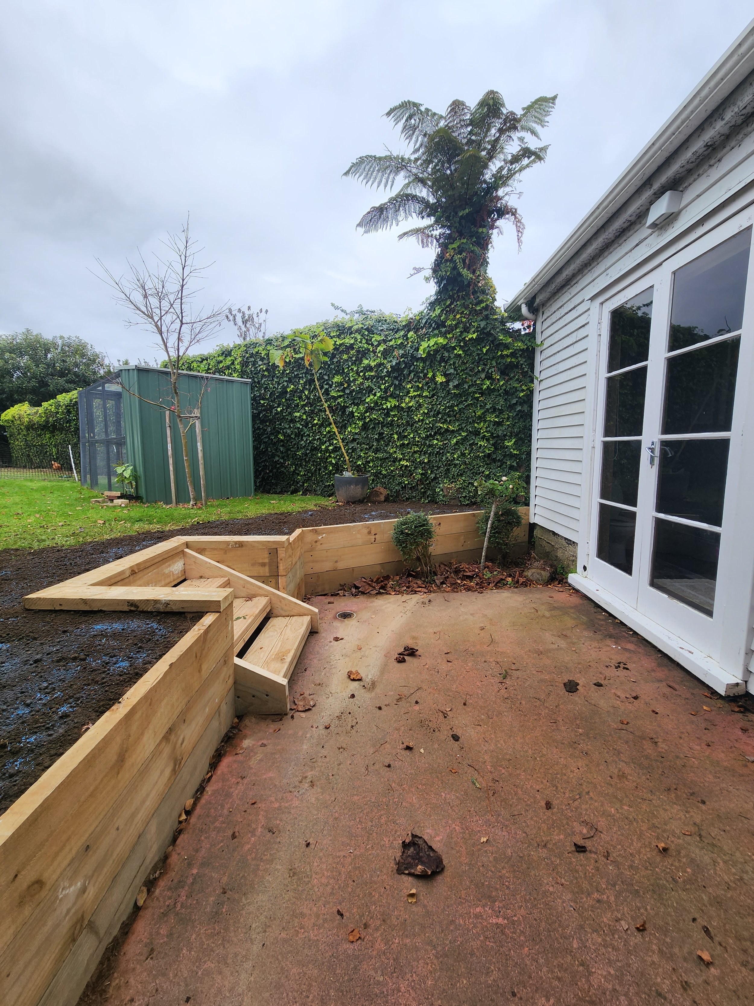 A backyard patio area with a newly built wooden raised garden bed and stairs, a hedge with climbing plants, a small potted plant, and a white house with glass double doors.