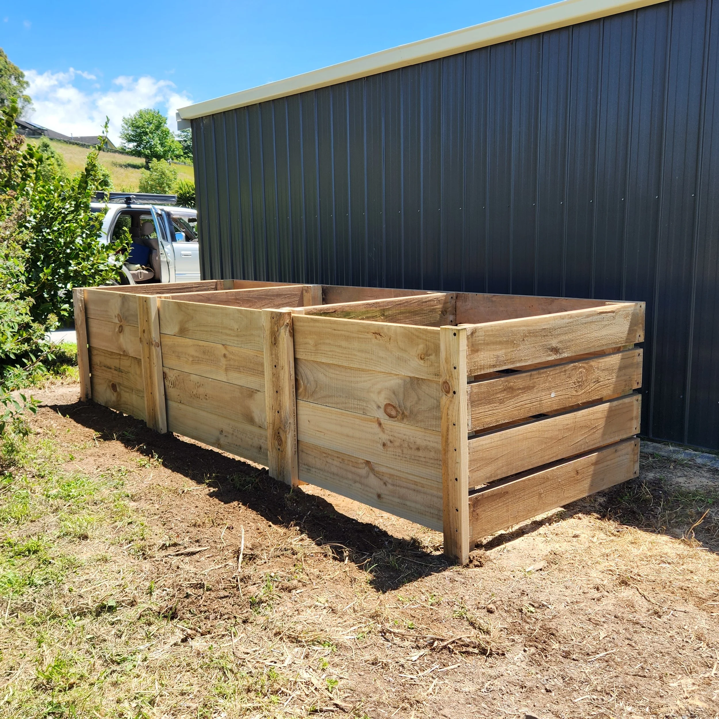 Newly built wooden garden bed with four sections, located beside a dark metal shed on a sunny day.
