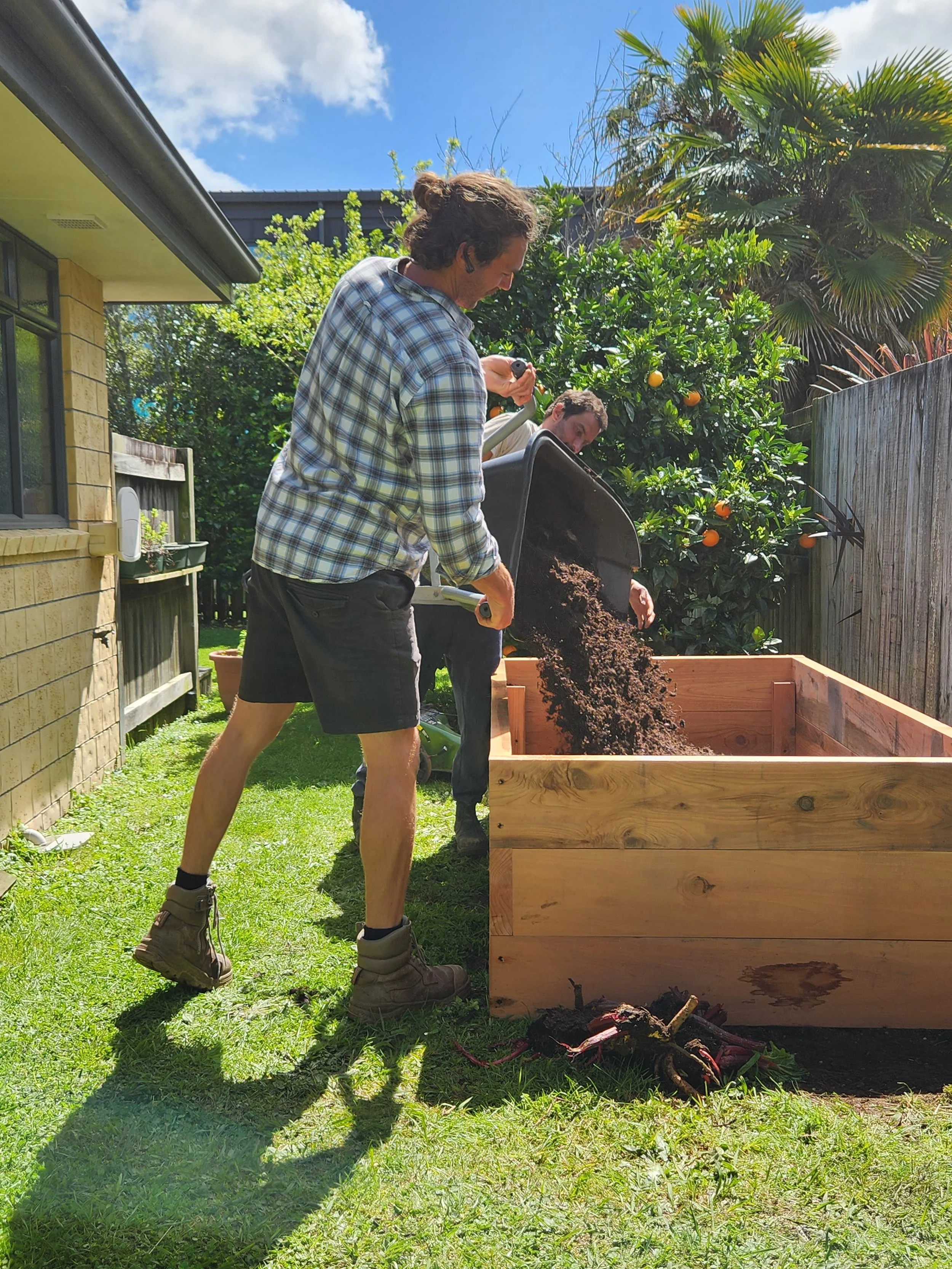 Two men fill a large wooden garden bed with soil in a backyard on a sunny day.