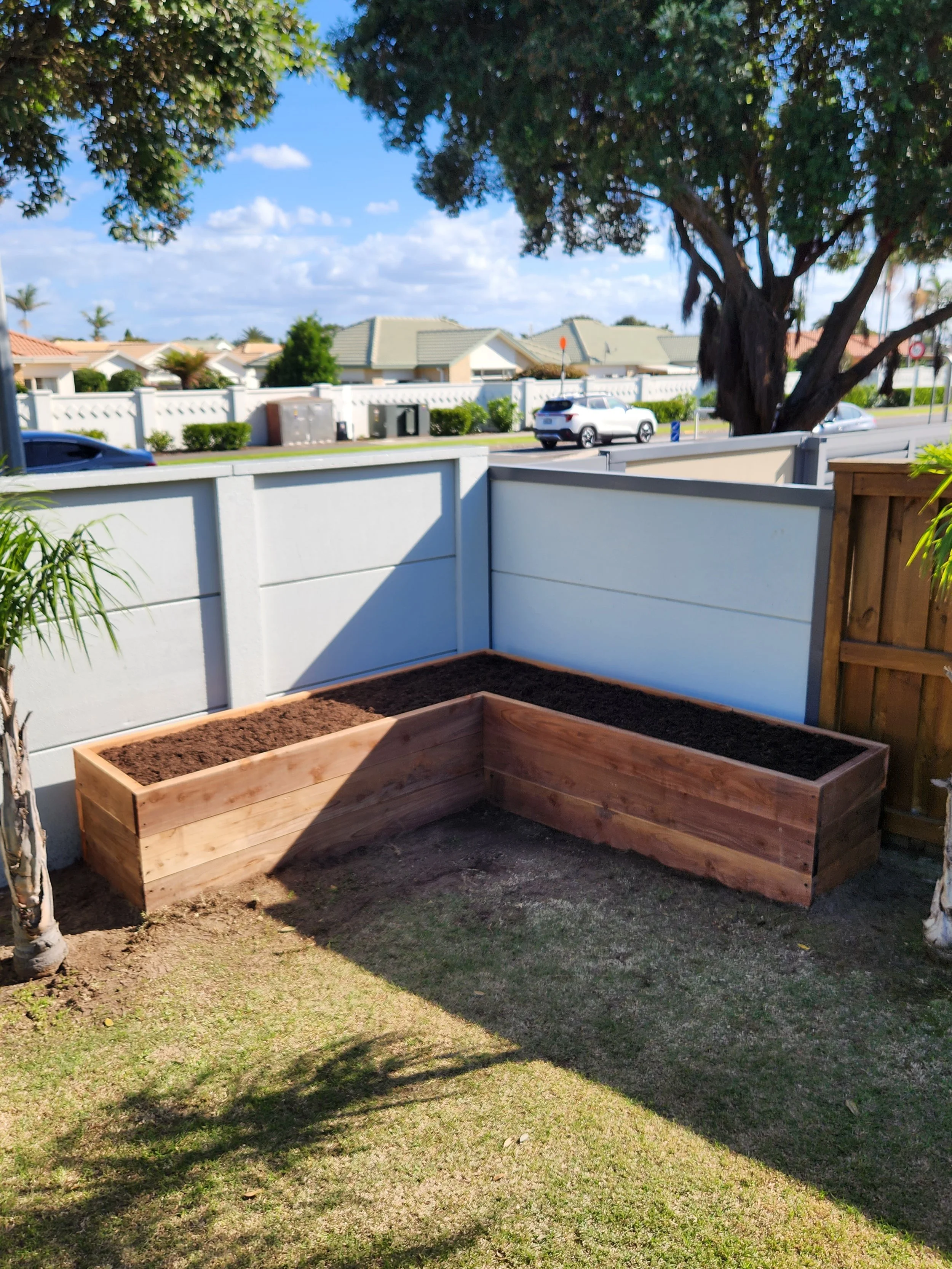 Empty wooden garden beds in a backyard with a white fence, green grass, and a large tree, under a blue sky with clouds.