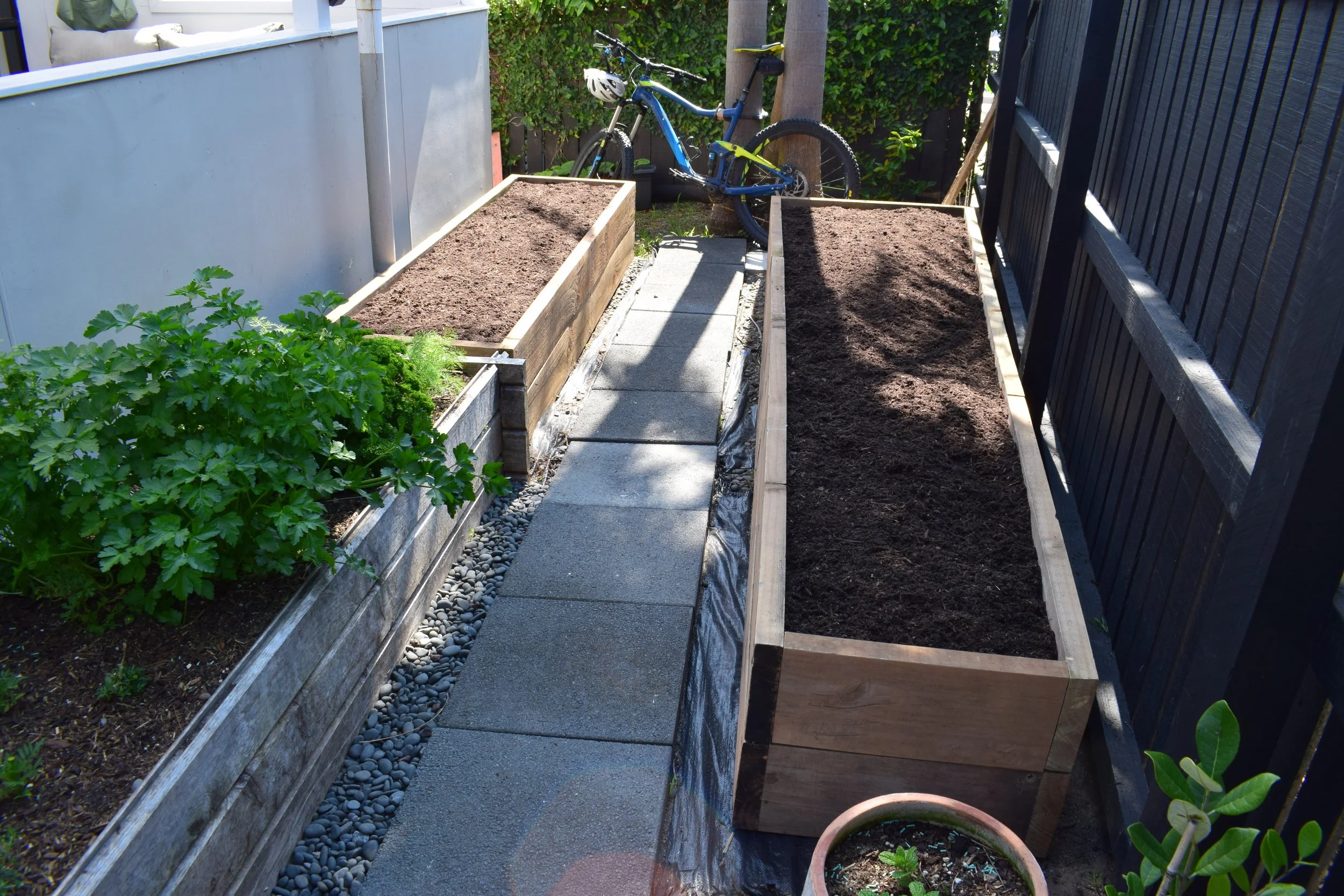 Garden beds with soil, a small potted plant, a dirt and gravel pathway, a bicycle, and a fence.