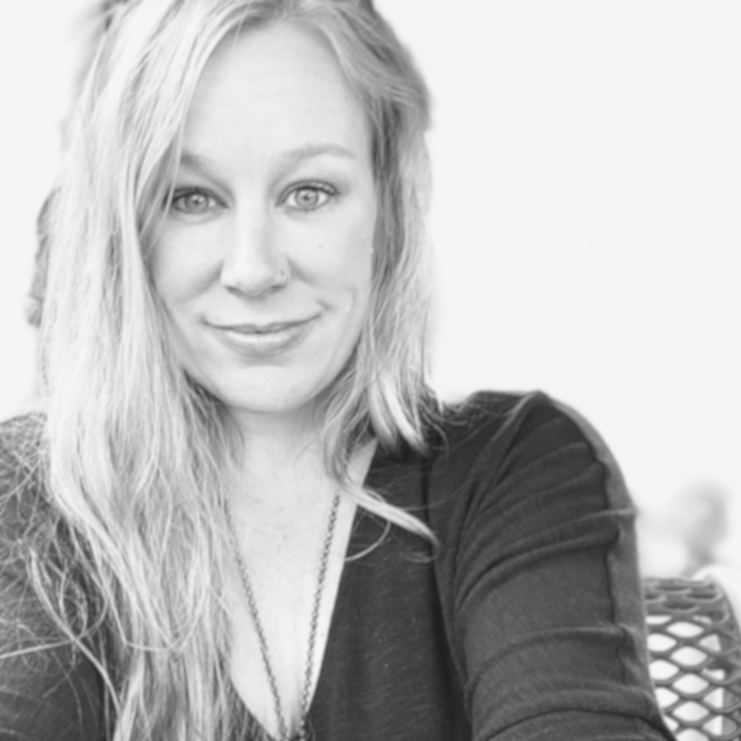A woman with long, wavy hair smiling and looking at the camera, wearing a dark top and a necklace, seated outdoors with a blurred background.