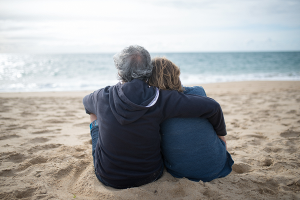 An elderly couple sitting on the sand at the beach, facing the ocean, with their arms around each other.