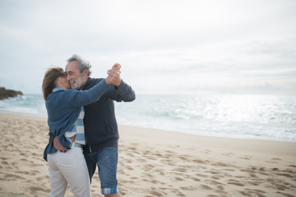 A happy couple dancing and kissing on a beach with the ocean in the background.