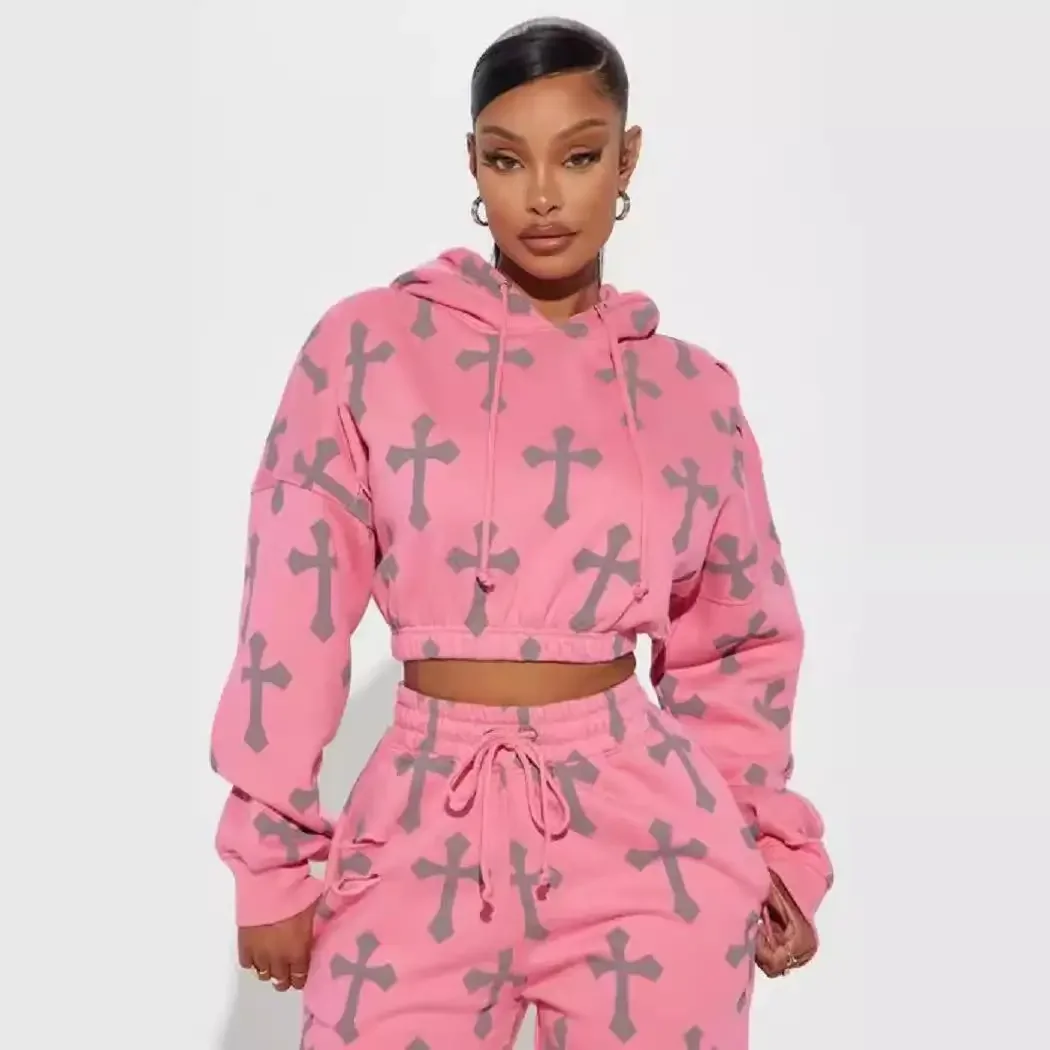 A woman with short black hair styled in a sleek look, wearing gold hoop earrings, poses against a plain white background. She is dressed in a matching pink hoodie and sweatpants, both adorned with a pattern of large gray crosses.