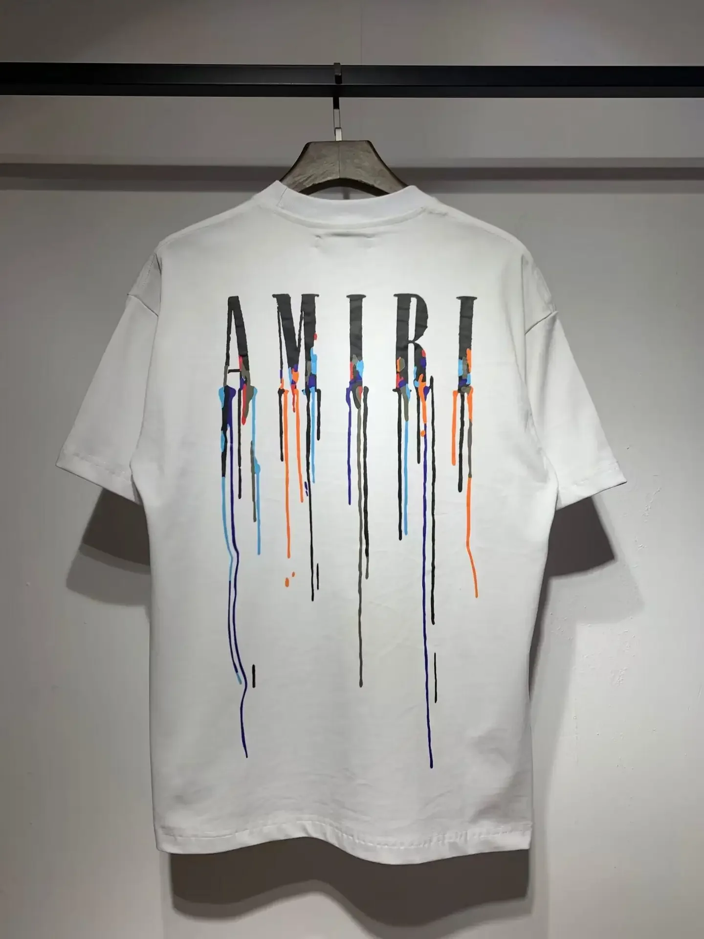 White t-shirt with the word 'AMIRI' printed on the back, featuring multicolored paint drips.