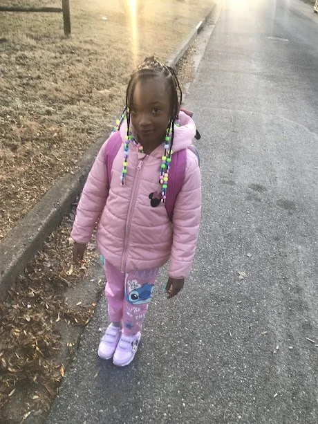 A young girl with beaded braids standing on a sidewalk wearing a pink jacket, patterned leggings, and light purple shoes.