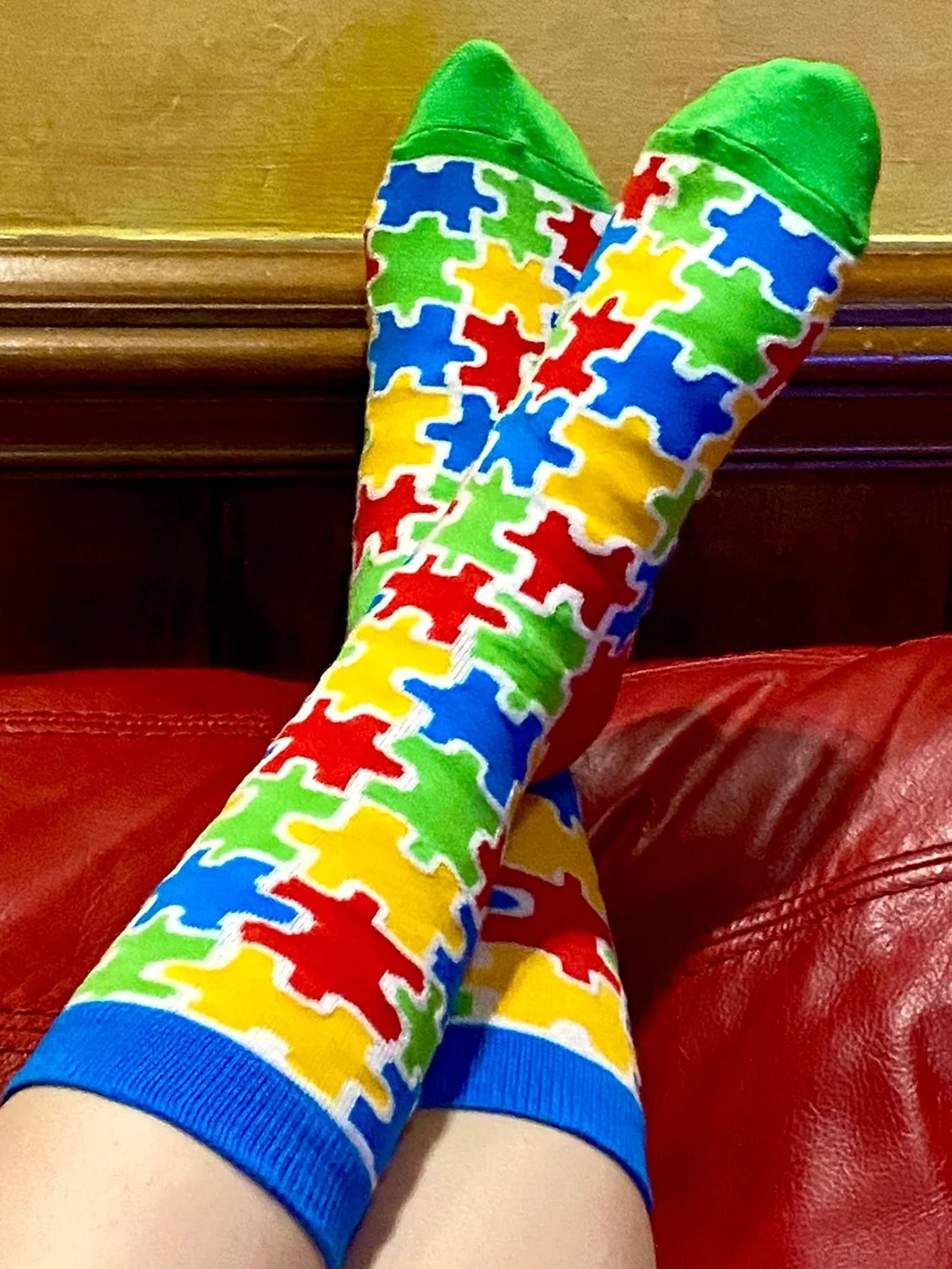 Colorful socks with puzzle piece pattern on someone's legs, resting on a red couch with a wooden headboard in the background.
