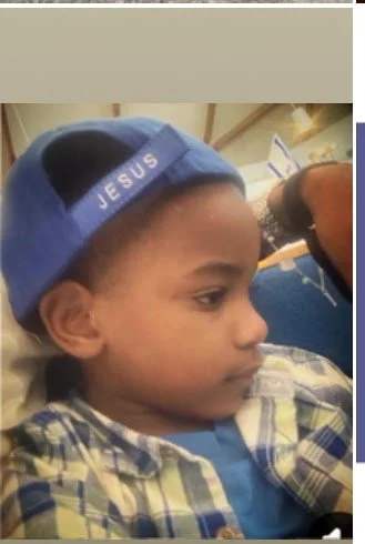 Young boy wearing a blue baseball cap with the word 'JESUS' stitched on it, sitting and looking to the side.