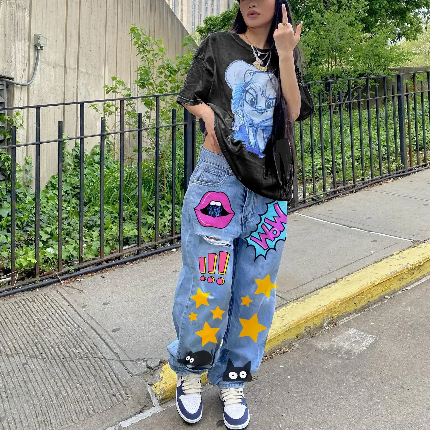 Person standing on sidewalk near a metal fence and greenery, wearing a black T-shirt with a cartoon character, loose jeans decorated with patches including a pink lips, exclamation marks, stars, a speech bubble with "WOW," and cartoon black cats, paired with sneakers.
