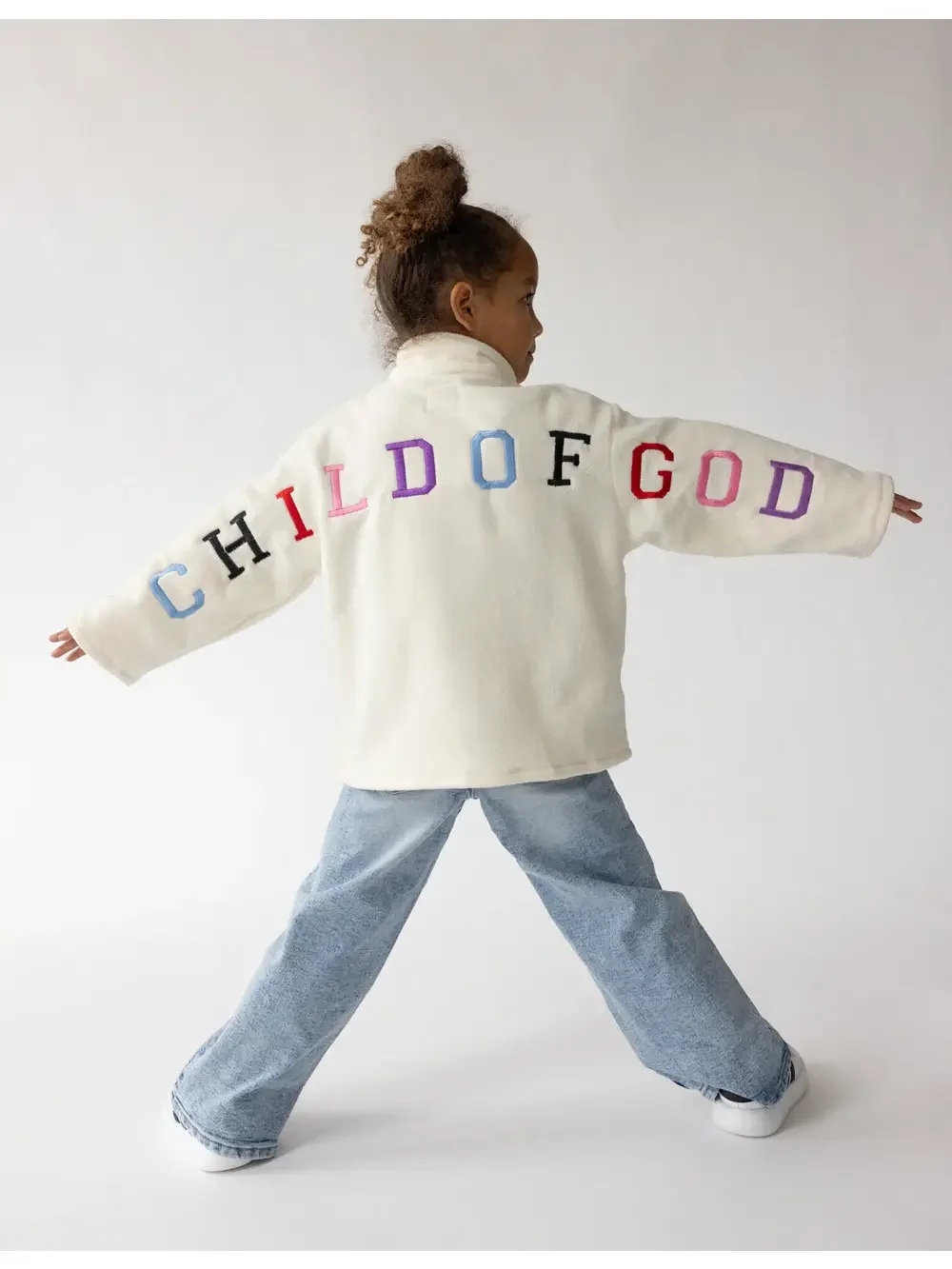 A young girl is standing with her arms outstretched, wearing a white jacket with the words 'CHILD OF GOD' embroidered in colorful letters across the back, light blue jeans, and white shoes.