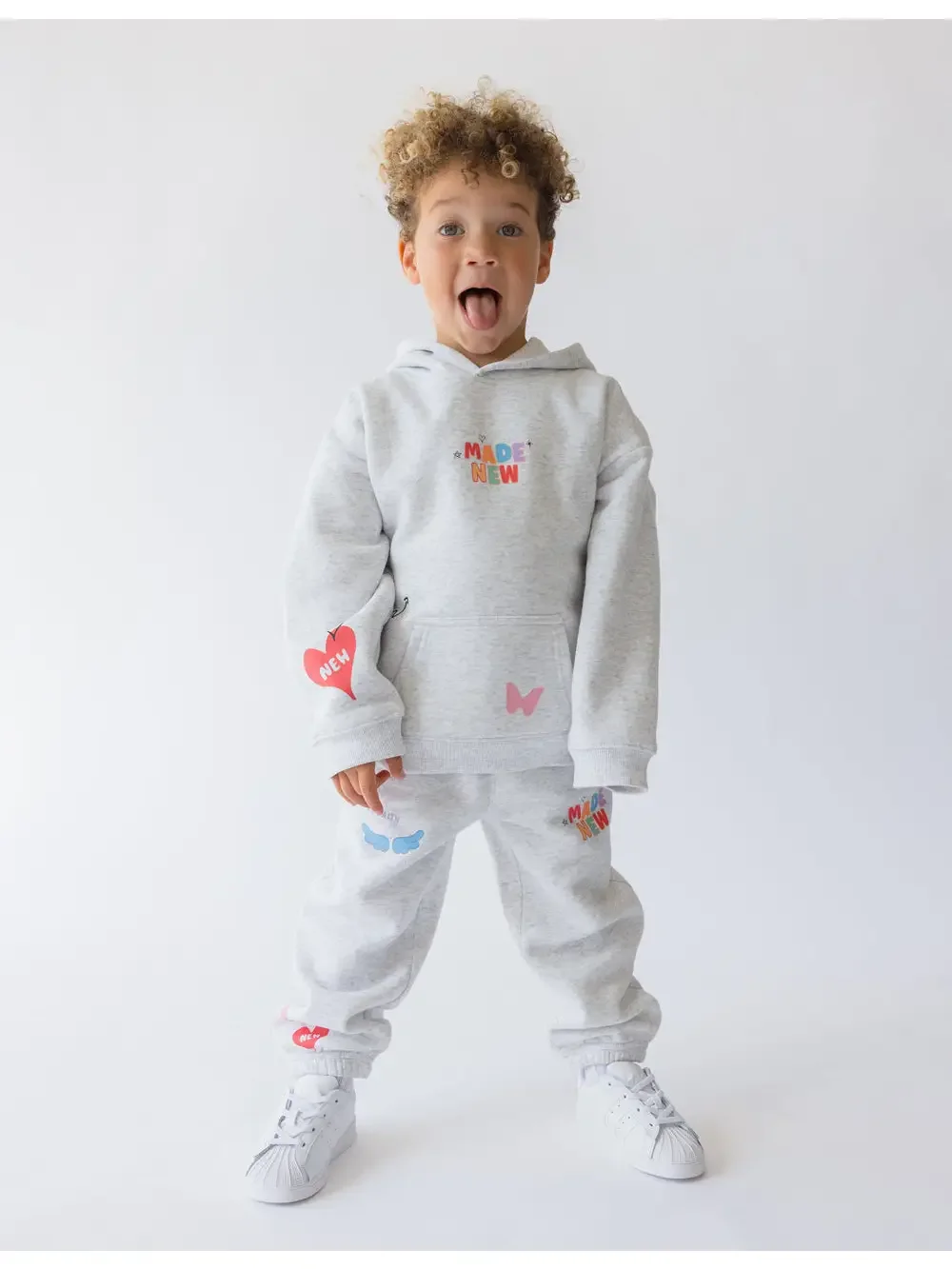A young boy with curly hair is standing and smiling with his mouth open, wearing a light gray hoodie and sweatpants decorated with colorful patches and words, along with white sneakers.