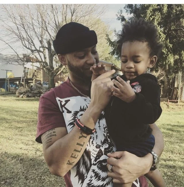 A man holding a young child outdoors, both looking at a smartphone. The man has tattoos and wears a cap and T-shirt with lion artwork. The child is touching the phone, appearing curious.