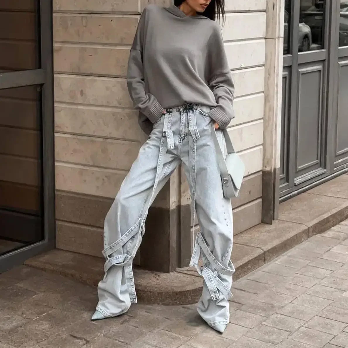 Person leaning against a wall wearing a loose gray sweater, oversized light-wash distressed jeans with long fabric ties, and white pointed shoes, holding a small white handbag, in an outdoor urban setting.
