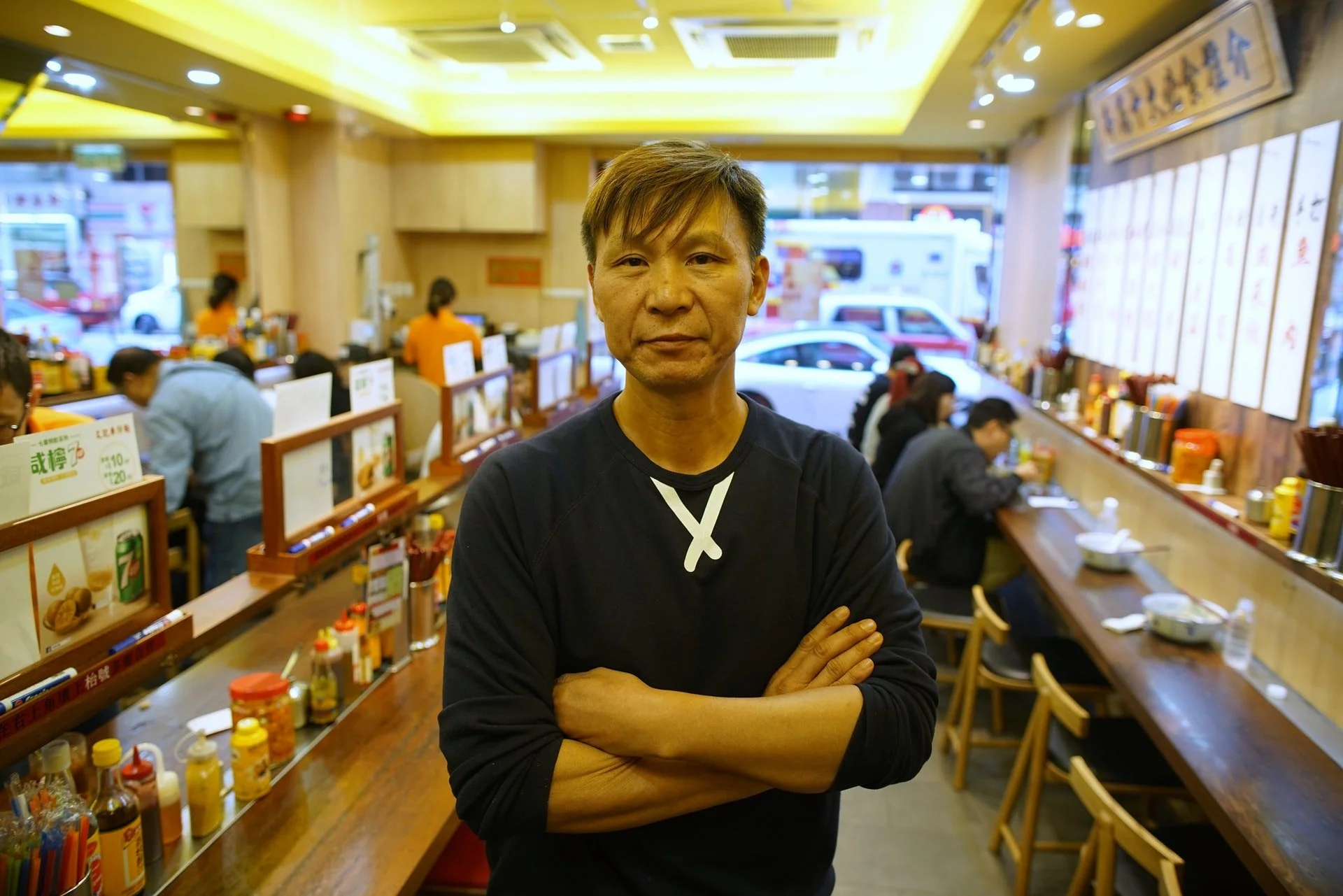 A man with crossed arms standing inside a busy Asian restaurant with a wooden counter, dishes, condiments, and diners in the background.
