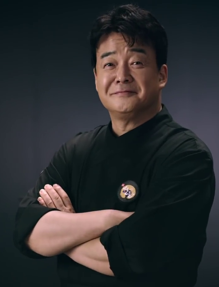 A man with dark hair, smiling with his arms crossed, wearing a black chef's coat with a badge, against a dark background.