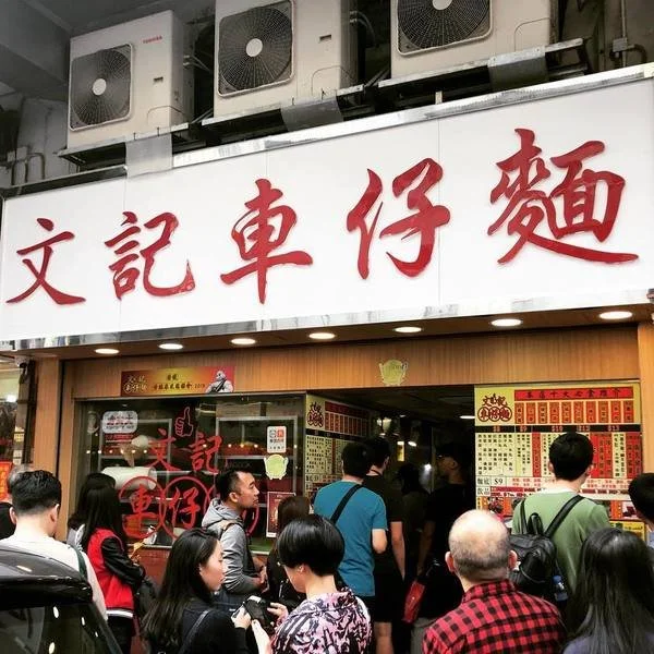 People waiting outside a Chinese restaurant with a sign in Chinese characters, featuring air conditioning units above.