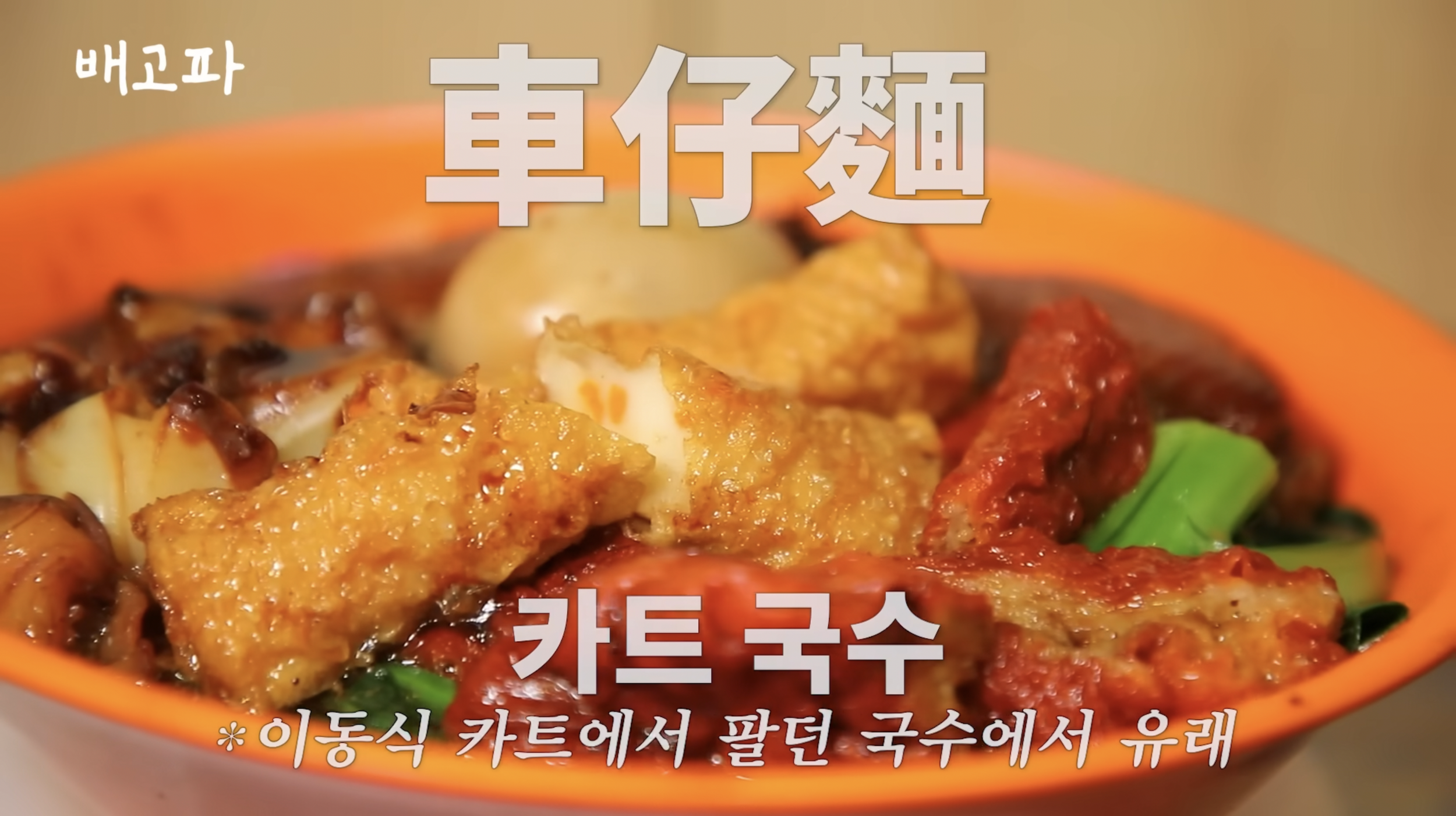 Close-up of a bowl of Korean vegetable noodle soup with fried chicken, mushrooms, vegetables, and red sauce, in an orange bowl.