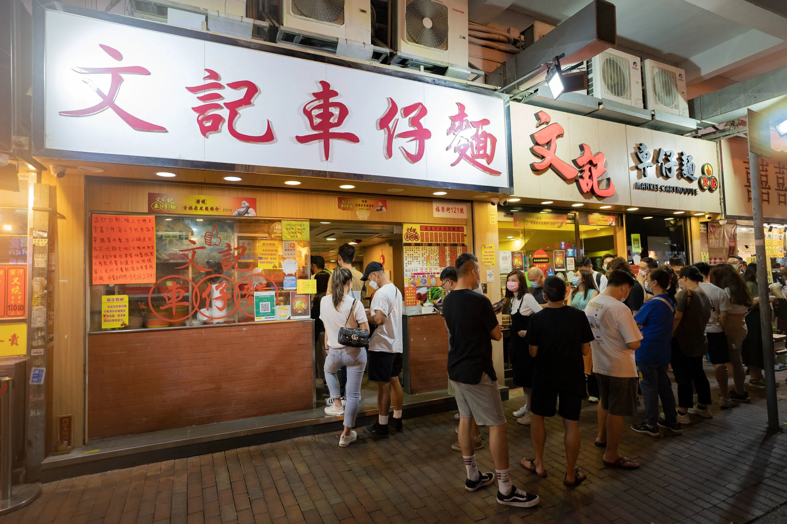 A crowded Asian restaurant with a red and white signboard displaying Chinese characters. People are waiting in line to order food inside.