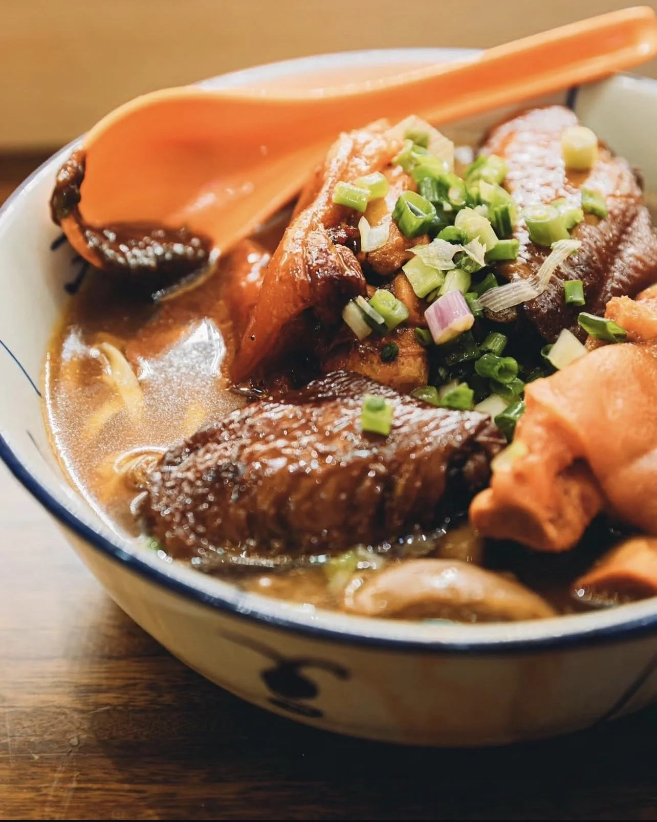 Bowl of Asian noodle soup with sliced pork, green onions, and broth. Orange spoon resting on the bowl's edge.