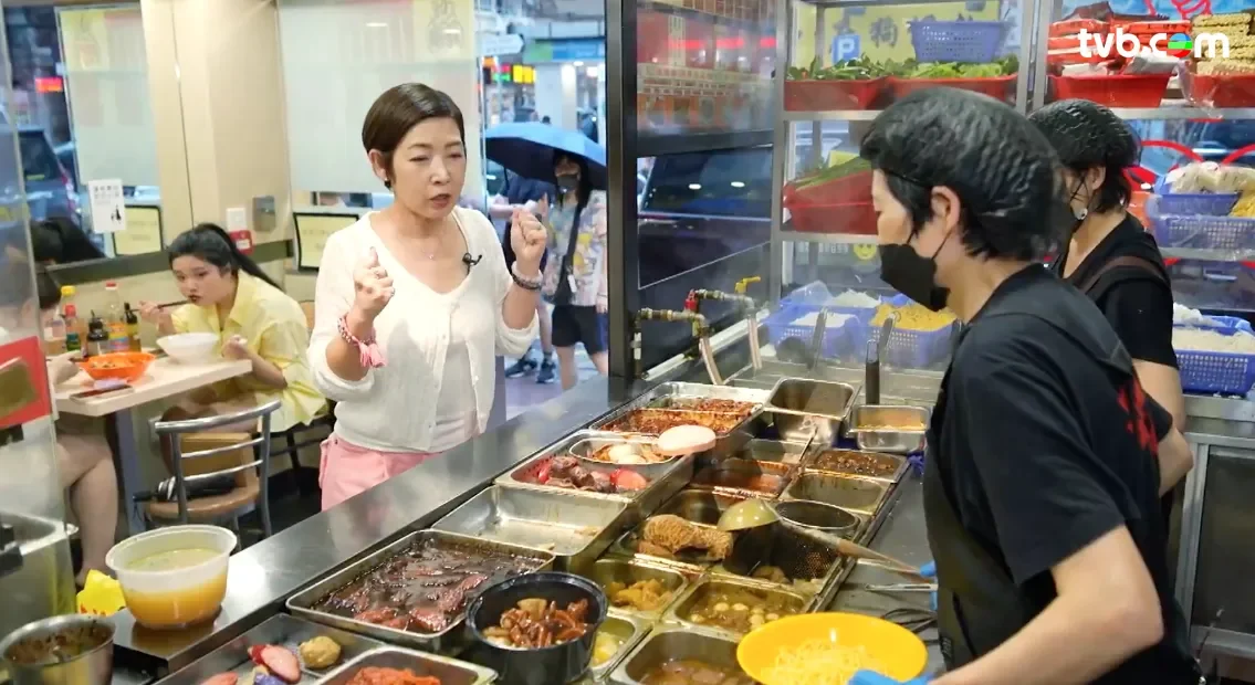 A woman talking to a worker at a food stall inside a restaurant, with customers dining in the background, some with umbrellas outside.