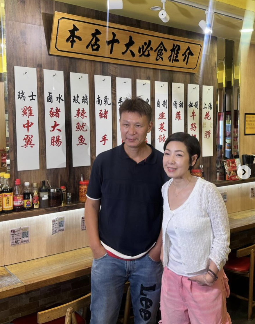 Two people standing inside a restaurant with Chinese menu signs on the wall behind them.