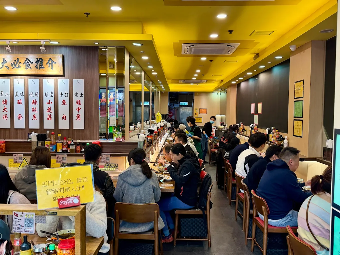 Busy Asian restaurant with many customers dining, some staff serving, and a counter area with condiments and menu signs on the wall.