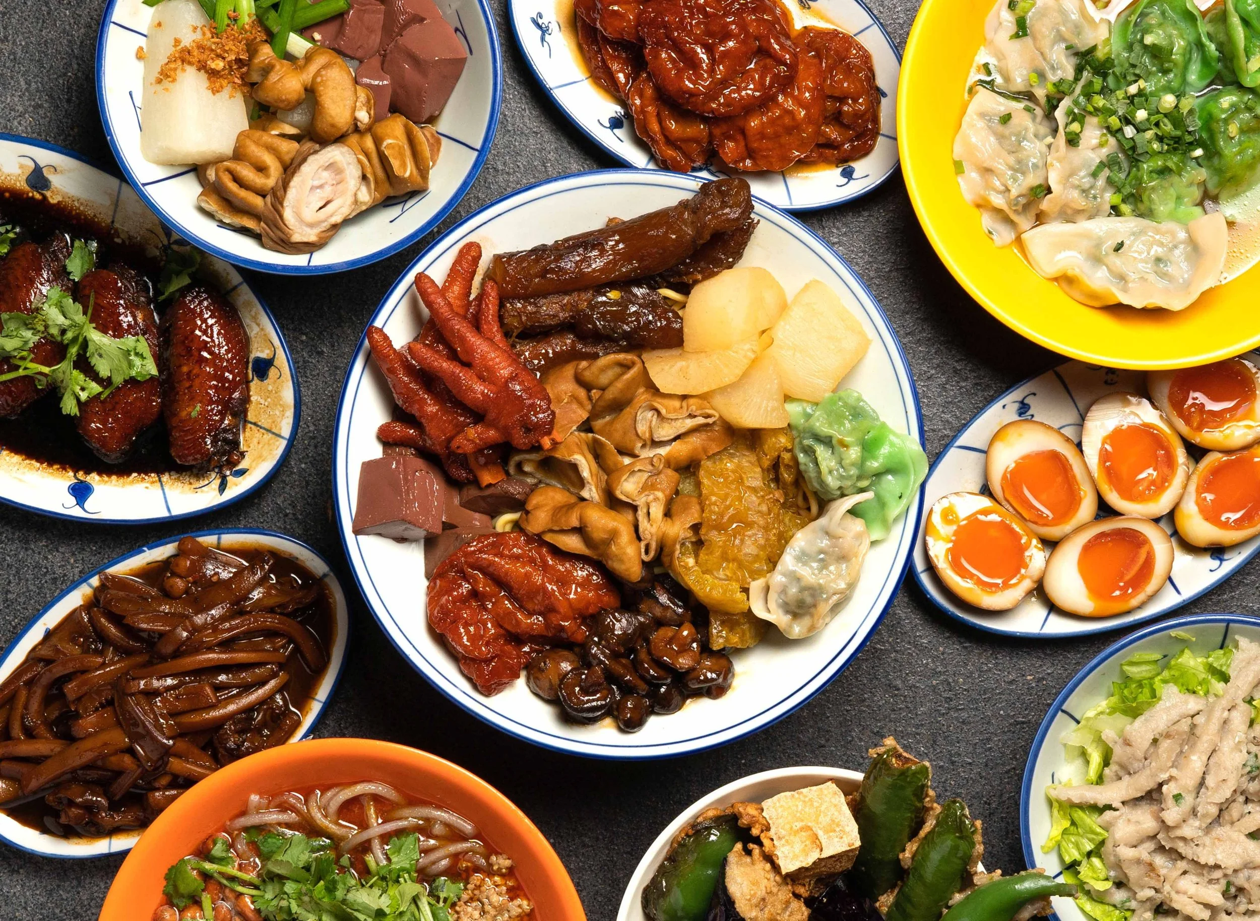 Assorted Chinese dishes served on small colorful plates, including dumplings, soft-boiled eggs, roasted meats, vegetables, and various sauces.