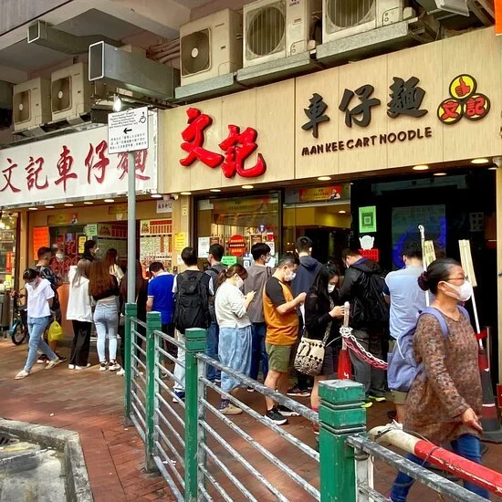 People waiting in line outside a restaurant named 'Man Kee Cart Noodle' in an urban area, with storefront signage in Chinese characters and English.