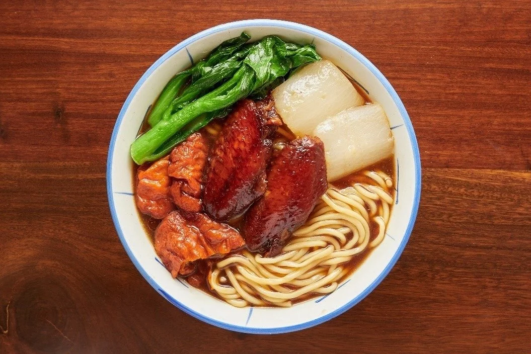 Bowl of Asian noodle soup with leafy greens, daikon radish, sliced cooked meat, and noodles in broth on a wooden table.