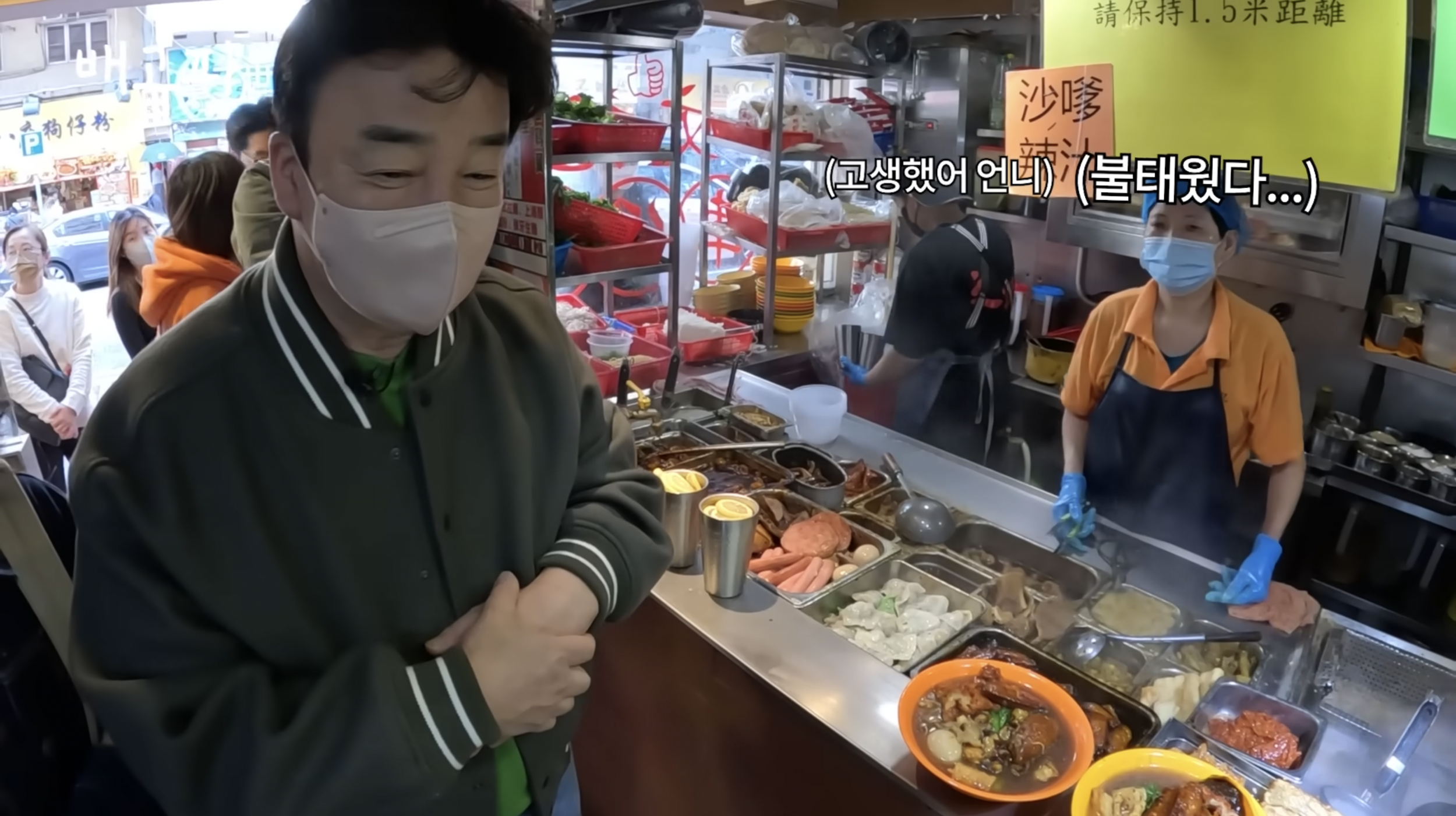 Man wearing a mask ordering at a street food stall with two women behind the counter, also wearing masks and gloves, serving hot dishes.