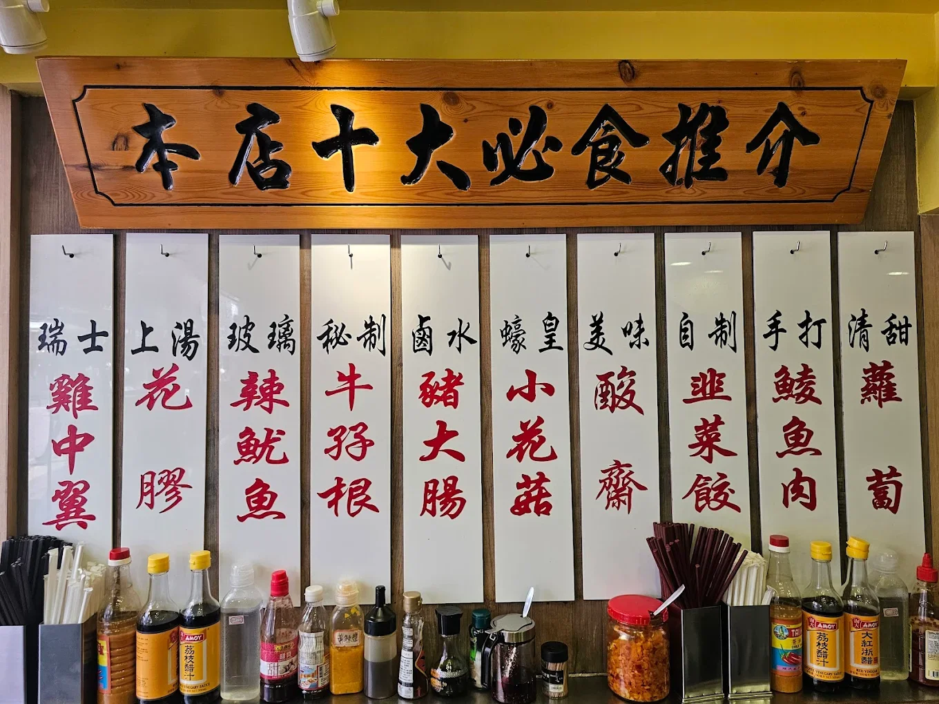 A wooden sign with Chinese characters, an assortment of condiment bottles, and containers with chopsticks and utensils on a restaurant counter.