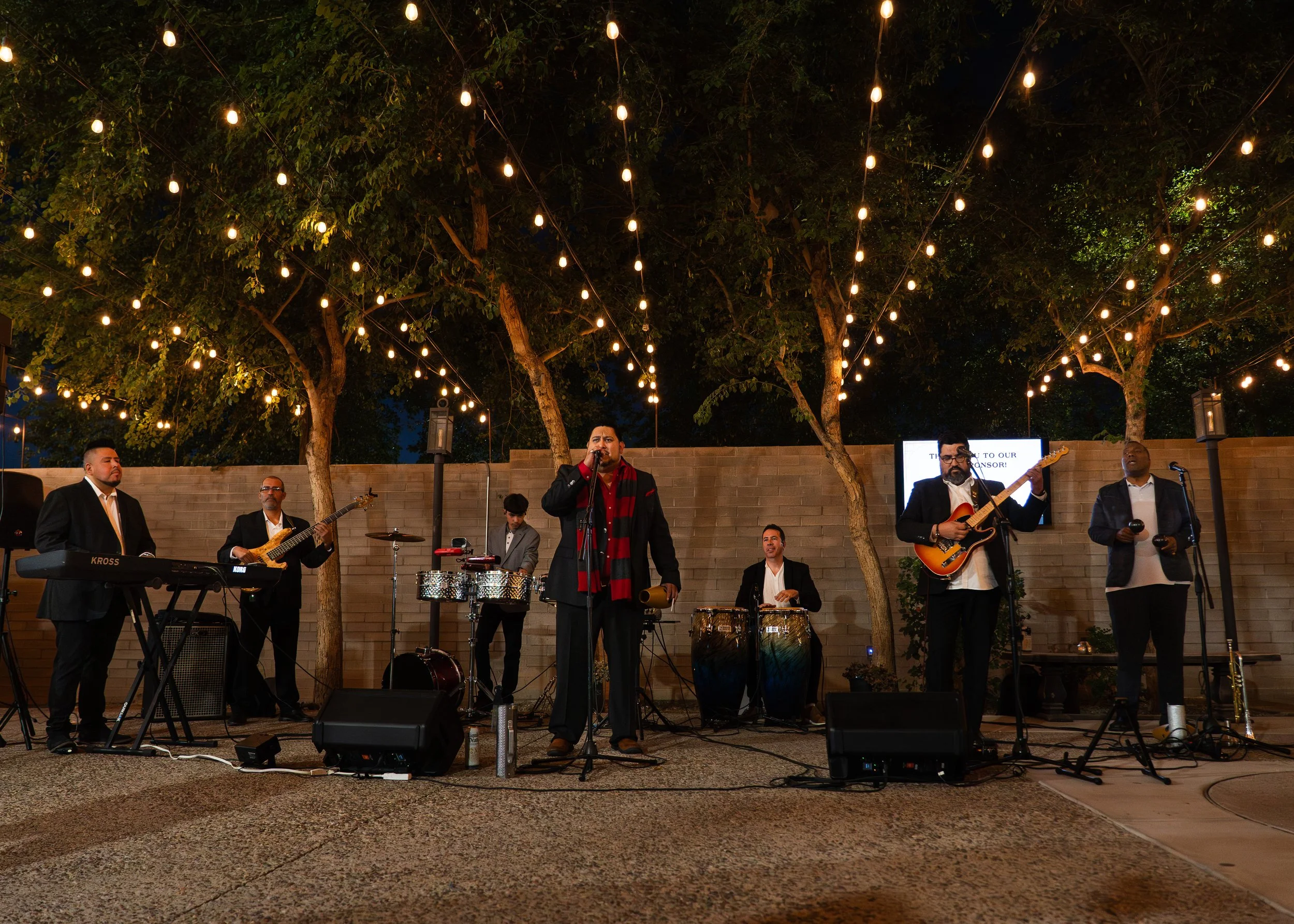 A live outdoor band performing at night under string lights, with six members playing various instruments including keyboard, bass guitar, percussion, and acoustic guitar, on a patio with a brick wall and trees in the background.