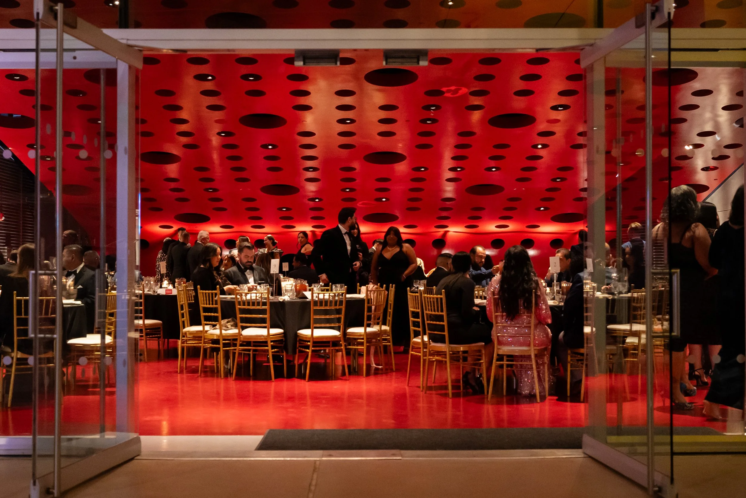 A lively indoor dining scene in a restaurant with a bold red wall featuring black circular patterns, gold chairs, and black tablecloths, with guests dining and waitstaff serving.