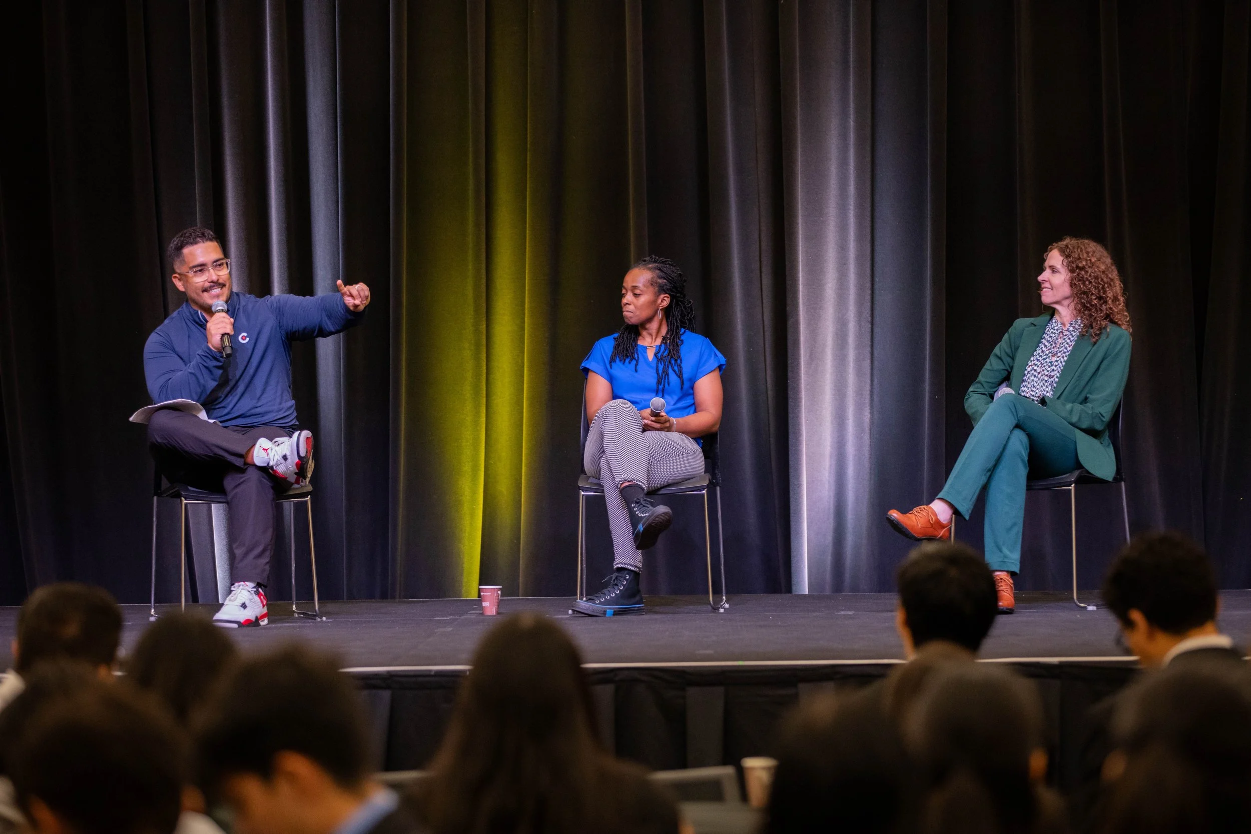 Three people sitting on stage in front of an audience, engaging in a panel discussion or talk, with dark curtains behind them.