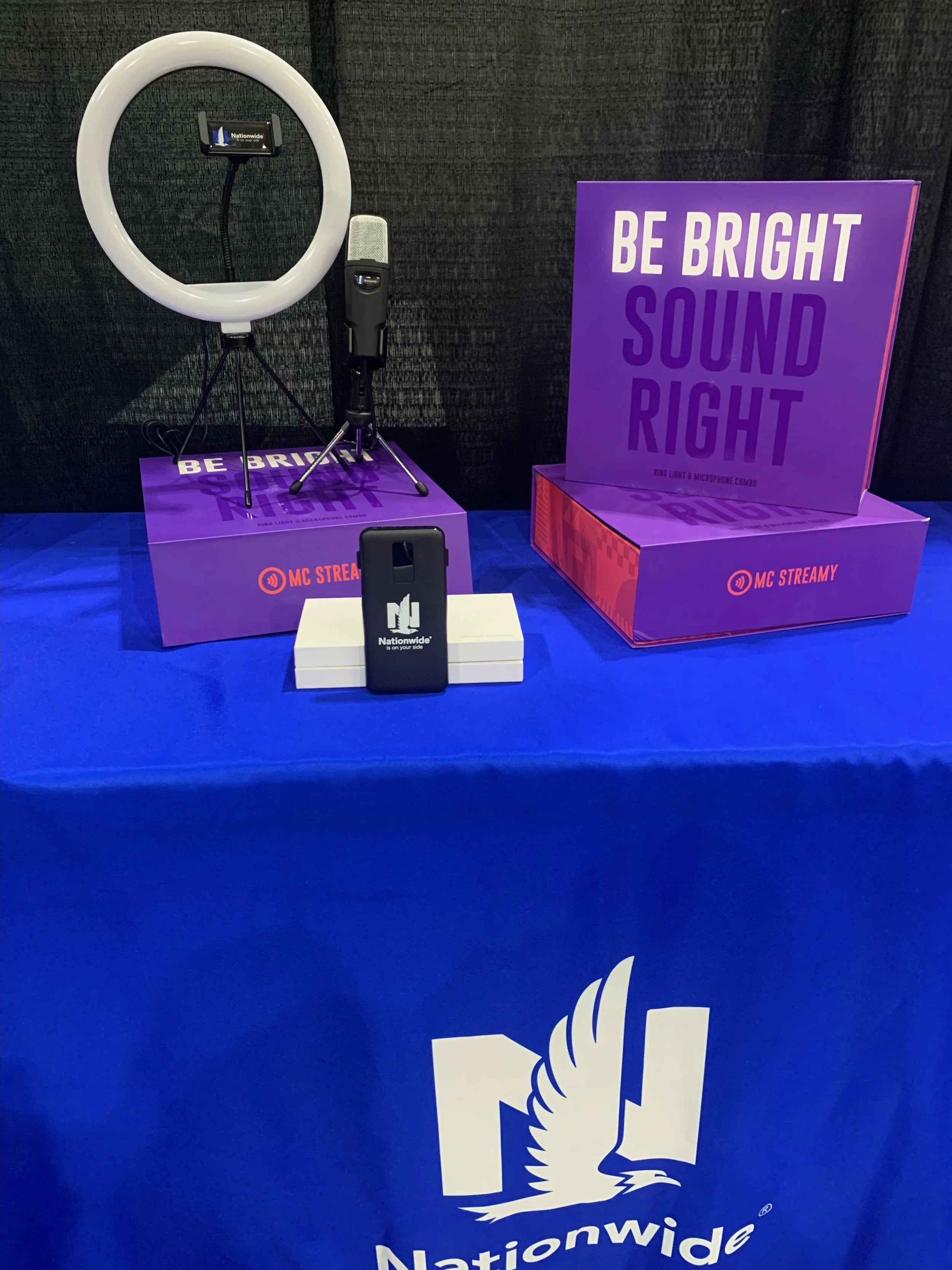 Display of a ring light, microphone, and smartphone on a table with a blue tablecloth featuring the Nationwide logo, and purple boxes with text "Be Bright Sound Right".